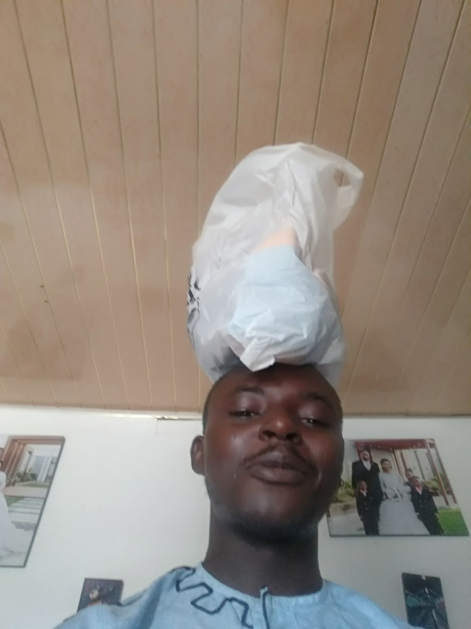 A man in Unknown, Unknown is standing indoors with a white plastic bag on his head. He is wearing a light blue shirt with a dark blue pattern around the neck. Behind him is a white wall with two framed pictures, one of a building and one of a family in formal wear. The ceiling is paneled in a light tan color.
