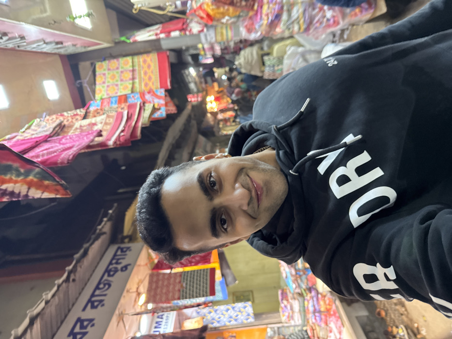 A young adult male, smiling directly at the camera, is centrally positioned in a close-up shot. He wears a black hooded sweatshirt with "DRY" in white capital letters across the chest and "24" on the right sleeve, along with a gold-colored chain around his neck. The background depicts a brightly lit, crowded market street at night in Kolkata, India. A shop sign written in Bengali script, partially visible on the left, reads "প্রবর রাজকুমার" (Probor Rajkumar). Stalls line the street, displaying an abundance of colorful textile products, including saris, patterned fabrics, and blankets, hanging from overhead racks and stacked on tables. Numerous out-of-focus individuals are visible in the background, indicating a busy commercial area.