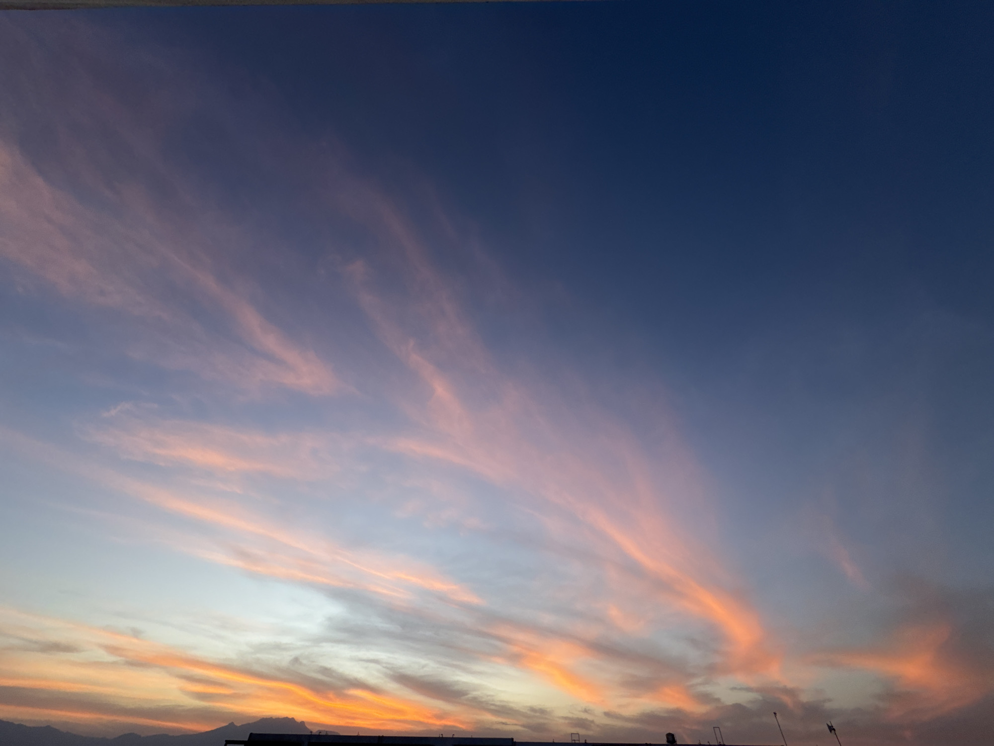 This media file captures a beautiful and expansive sky scene, most likely at sunset or sunrise, over San Francisco Ocotlán, Mexico. The dominant subject is the sky, featuring a captivating gradient of colors. The upper portion of the sky is a deep, rich indigo blue, gradually fading into lighter shades of blue and purple towards the middle.
Below this, wispy, elongated clouds are illuminated with warm, vibrant hues. These clouds are predominantly pink, orange, and peach, appearing in streaky formations that stretch across the frame, especially prominent on the left side where they seem to radiate upwards. The intensity of these warm colors is strongest in the lower half of the sky, blending into lighter, softer tones.
At the very bottom of the image, a dark, silhouetted horizon line is visible. On the left, this silhouette suggests distant mountains or hills. On the right, faint dark shapes indicate structures, possibly rooftops or buildings with antennas and poles, against the glowing sky. A very thin, dark line, possibly a wire or edge of an overhead structure, runs horizontally along the very top edge of the frame.
The overall scene conveys a sense of tranquility and natural beauty, characteristic of dawn or dusk. There are no visible people or discernible text. The weather appears clear with high-altitude cloud formations, contributing to the dramatic sky display.