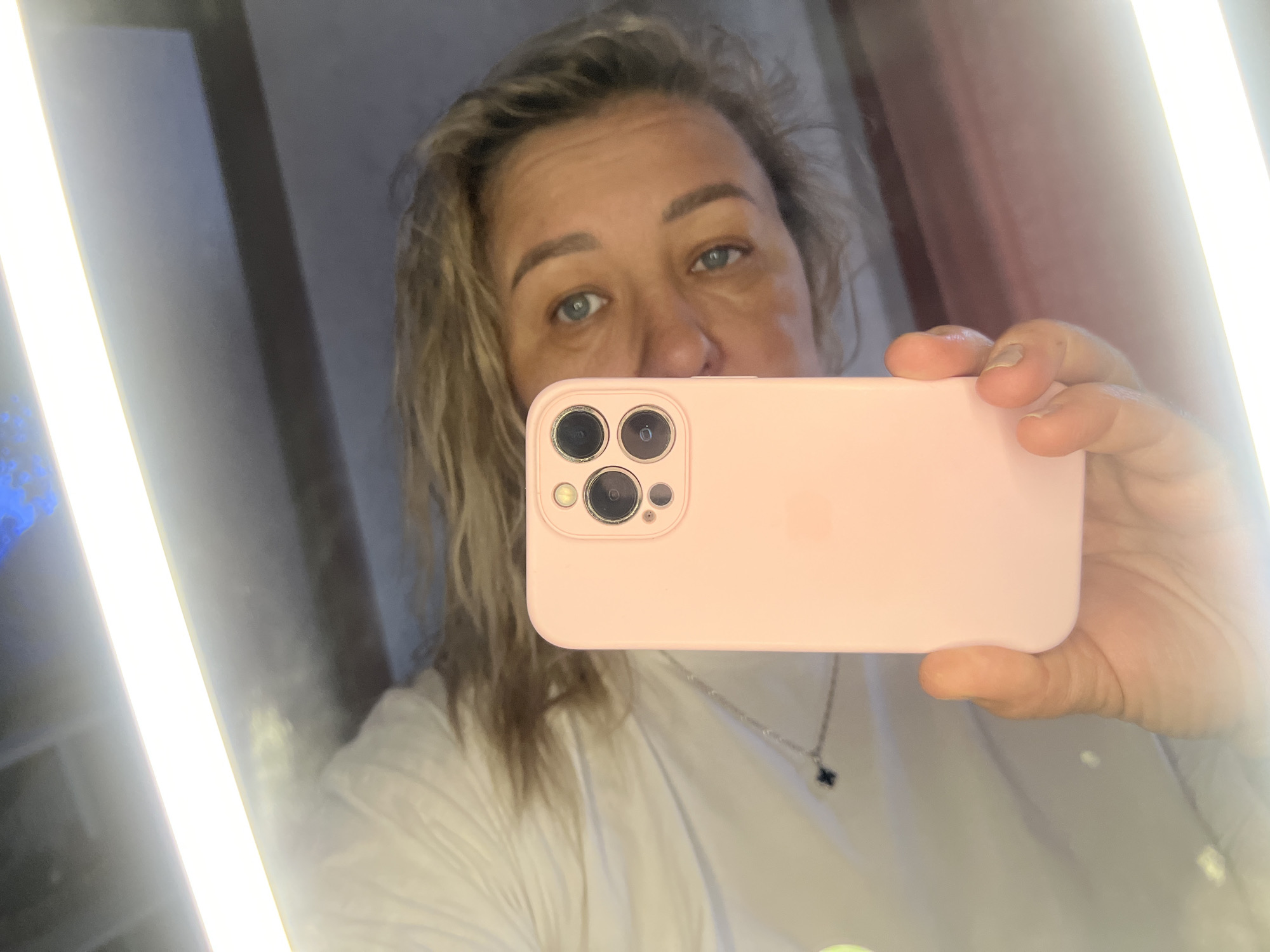 An adult female is captured in a mirror selfie, holding a pink smartphone that partially obscures her face. Her light-colored hair is visible around her face, and her blue-green eyes look directly towards the phone's lens. She appears to be wearing a light-colored, possibly white, t-shirt and a delicate silver necklace with a dark, four-leaf clover-like charm. Her right hand holds the smartphone, which features a triple-lens camera system on its back.
The setting appears to be an indoor space, likely a bathroom or dressing area, characterized by two bright, vertical light strips flanking the mirror, providing illumination. These lights are prominently visible on both sides of the reflection. Hints of a darker background, possibly a wall or curtain, can be seen beyond the light strips. The image captures a moment of self-portraiture within an illuminated interior in Chaikovsky, Russia. There is no visible text in the image.