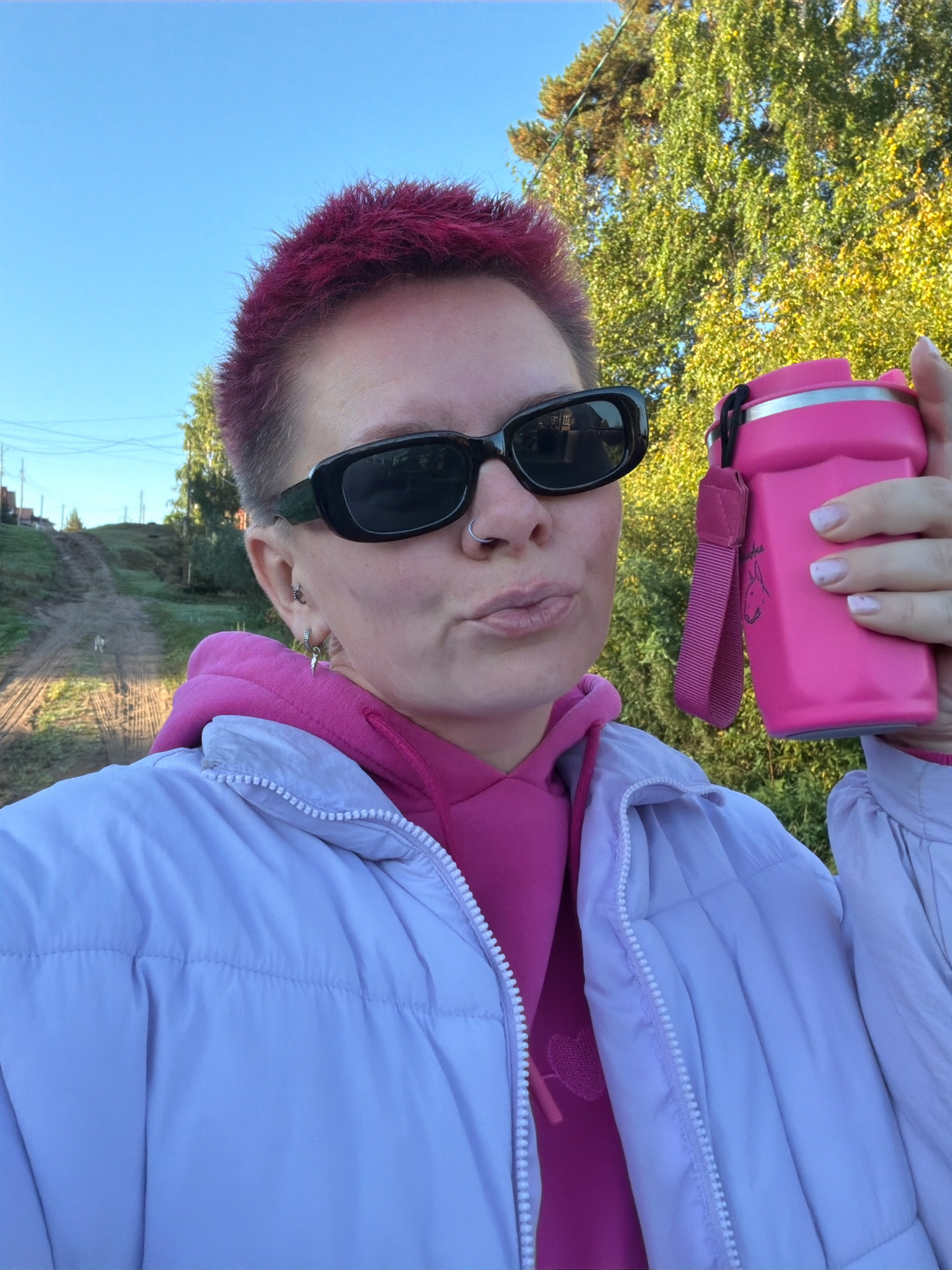 A person with short, magenta-dyed hair and black sunglasses takes a selfie outdoors. They are wearing a bright pink hoodie and a light purple puffer jacket. In their right hand, they hold a pink water bottle with a dark pink strap and a small cartoon drawing of an animal. The background shows a rural setting with a dirt road leading uphill, trees with yellowing leaves, and a clear blue sky. The location is identifiable as Chaikovsky, Russia.