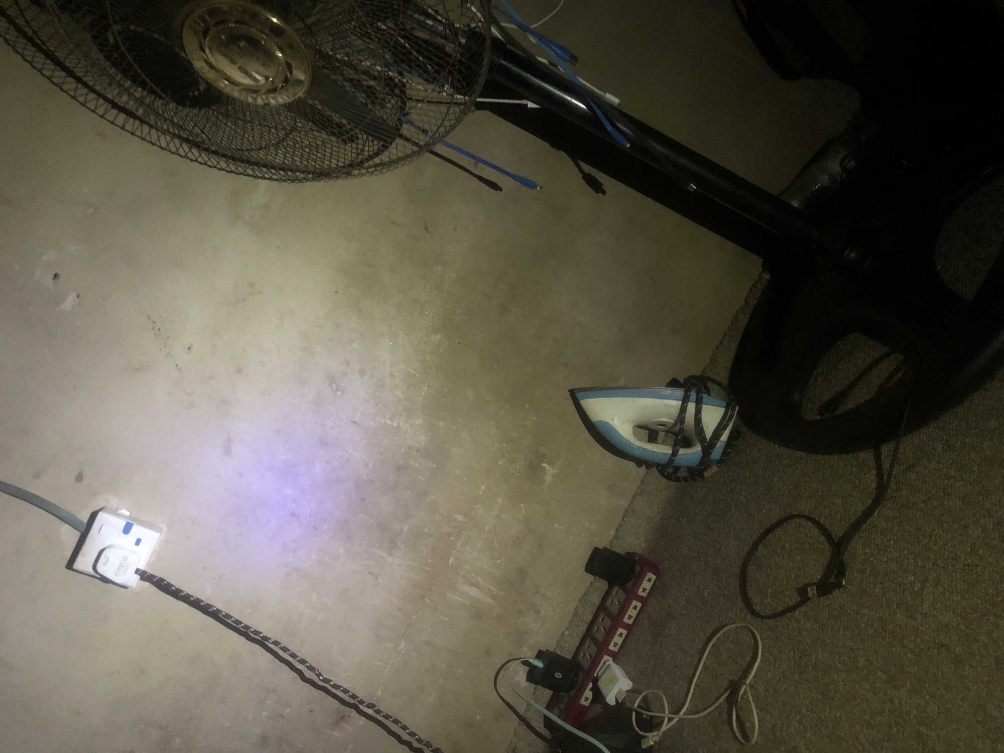 The image depicts an indoor scene with various electrical appliances and accessories. In the upper left, a white electrical socket is mounted on a textured, light-colored wall, with a black cord plugged into it. A purple light emanates from the socket area. Below this, a red power strip with multiple outlets is visible, with several devices and cords connected to it. To the right of the power strip, a blue and white electric iron is resting on its heel. In the lower right, the base and lower portion of a black standing fan are present, with blue cords hanging from its cage. The floor appears to be carpeted with a dark, textured material. The lighting is dim, creating shadows and highlighting certain objects. The environment suggests a domestic or workspace setting, possibly a storage area or a room where electrical devices are in use or being charged. The image is not set in the city of Jalingo, Nigeria, and no information about that location is present or inferable from the visual content.