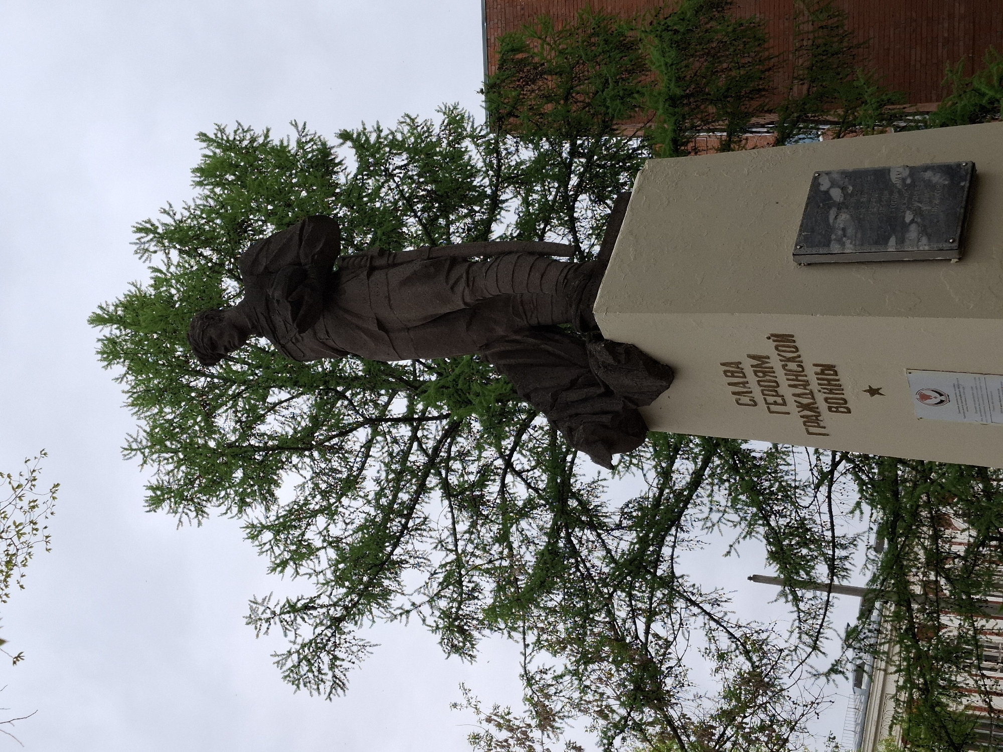 The image depicts a monument to the heroes of the Civil War in Glazov, Russia. The monument consists of a bronze statue of a soldier on top of a concrete pedestal. The pedestal has inscriptions in Russian and a plaque with images. The monument is surrounded by trees, and the sky is overcast. The monument appears to be located in a park or public square.