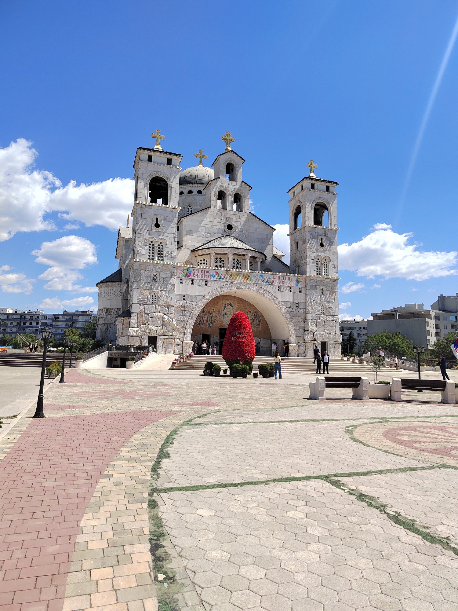 The image depicts the Cathedral of the Resurrection of Christ in Podgorica, Montenegro, on a sunny day with scattered clouds. The cathedral, constructed of light-colored stone, features two prominent bell towers, a central dome, and multiple golden crosses. Banners adorn the entrance arch. A large, cone-shaped, red floral arrangement stands in front of the cathedral entrance, partially obscuring the stairs and entrance. Several people are visible near the entrance, some on the steps and others standing on the plaza. The plaza is paved with patterned stone, with sections of red brick walkways. Benches and lampposts are spaced across the plaza. In the background, buildings are visible beyond the plaza, indicating an urban setting. A street is seen on the left side of the image.