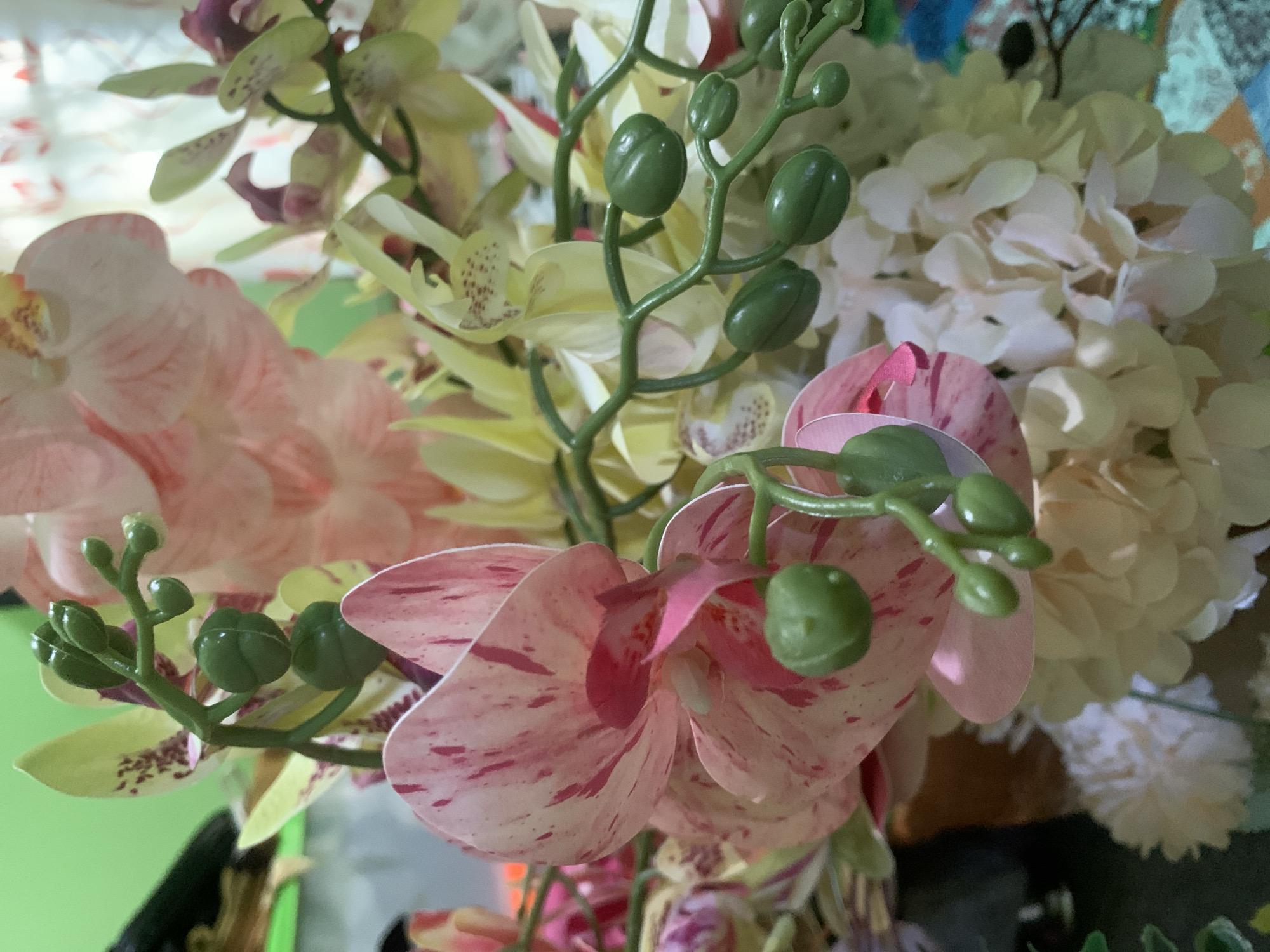 This is a close-up, eye-level, vertical shot of a bouquet of artificial orchids and other flowers. The bouquet is primarily composed of pink and white orchids, with some pale yellow and green accents from other artificial flowers and the orchid stems and buds. The lighting is moderate, with some areas appearing slightly overexposed, particularly in the background. The background itself is blurred and out of focus, showing hints of a patterned surface with blues, oranges, and possibly some floral elements. There are no people or animals present in the image. The scene appears to be indoors, possibly on a table or shelf, given the out-of-focus background which could be fabric or a decorated surface. The image is too focused on the artificial flowers to provide any context about the location within Kuala Pembuang, Indonesia.