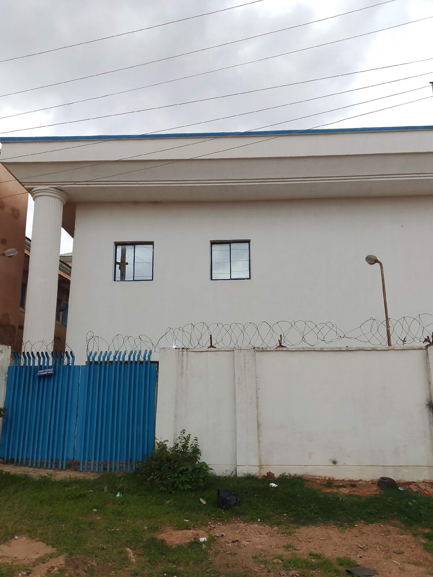 The image shows a white building with a blue trim on top, under a cloudy sky. The building has a large white column on the left side. There are two dark-framed windows on the building's facade. In front of the building is a tall, bright blue metal gate. Above the gate, a white concrete wall is topped with coiled barbed wire. A rusty streetlight stands to the right of the building. The ground in front of the wall is covered with grass and some debris, including plastic bags and bottles. A sign on the blue gate reads "CARS PARKED AT OWNER'S RISK."