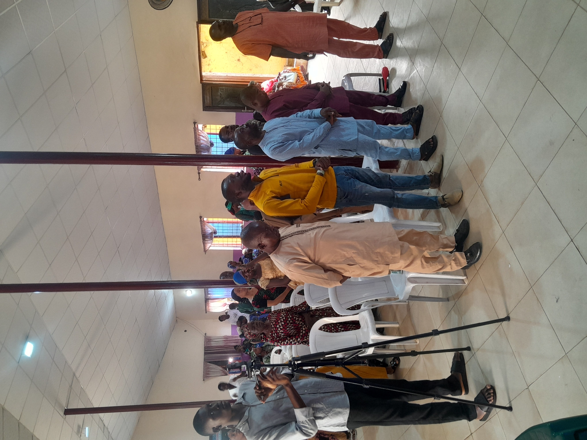 The image shows a group of people sitting in rows in what appears to be a church or large meeting hall in Jos, Nigeria. A man in the foreground is holding a camera or phone mounted on a tripod. The people are of African descent. The room has a tiled floor, a white tiled ceiling, and dark poles in the middle. Windows are visible along the walls. The people are sitting on white plastic chairs. The subjects appear to be sitting and facing forward. Some people are in traditional African clothing.