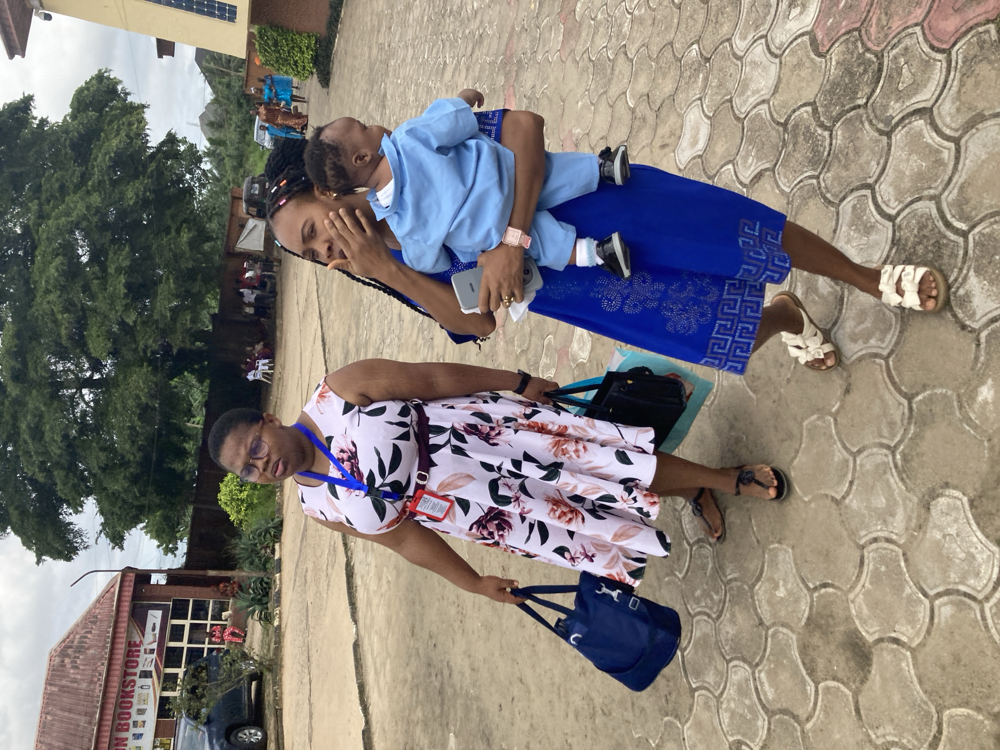 In Umuode, Nigeria, two women and a baby are visible outdoors on a cloudy day. The woman on the left, wearing glasses and a floral dress, carries two bags, one dark blue and the other black. She stands on patterned paving stones. To her right, a woman in a blue dress holds a baby wearing a light blue outfit. She is also on the patterned paving stones. In the background, a building with the sign "ON BOOKSTORE" is visible, along with a dark blue car parked in front of it. Trees and other people are also present in the background.