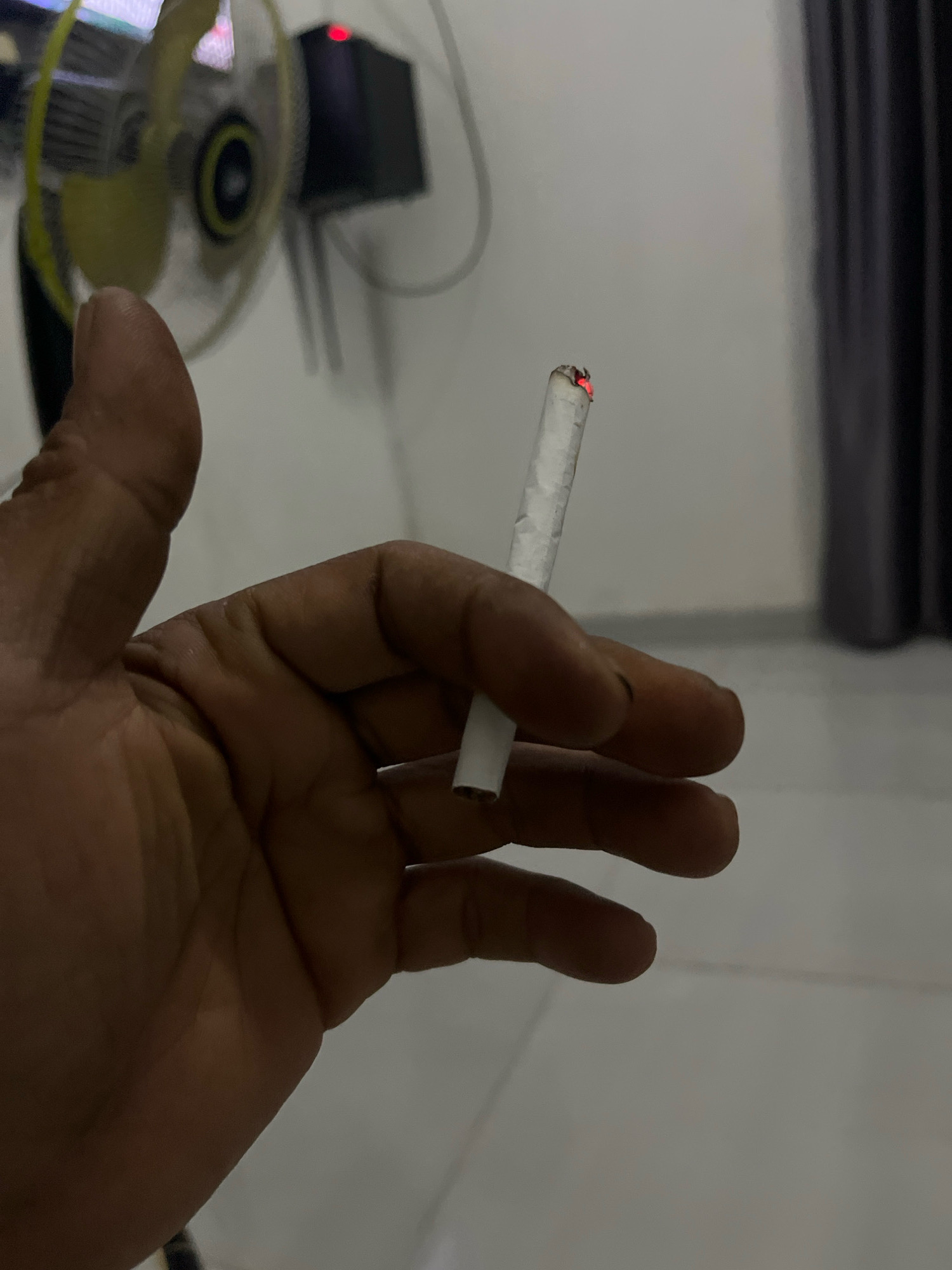 A close-up shot shows a person's hand holding a lit cigarette. The hand is dark-skinned, with visible wrinkles and fingernails. The cigarette is white with a red glowing tip, indicating it is burning. In the blurred background, a yellow fan and a black object with a red light are visible. The setting appears to be indoors with a light-colored wall. There is no specific information to determine the location in Kebumen, Indonesia.