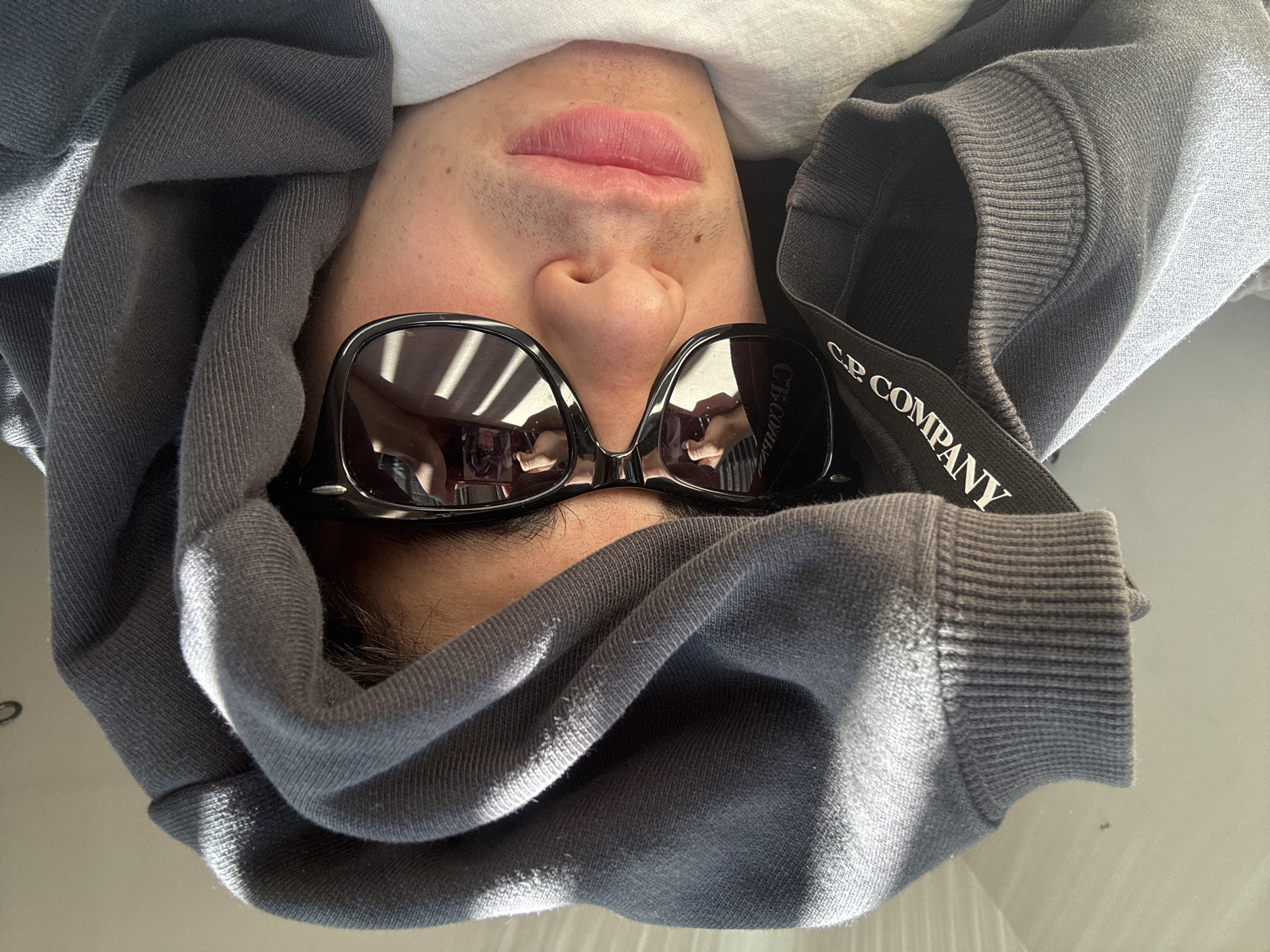A person with light skin is facing the camera while lying on their back. They are wearing sunglasses that reflect a room with light-colored walls and dark furniture or window frames. The person's mouth, nose, and a bit of stubble are visible below the sunglasses. They are wearing a gray hooded sweatshirt, with the hood framing their face. Part of the hood has a black strap visible that reads "C.P. COMPANY" in white lettering. Above the hood is a small area of a white fabric. The background consists of a plain surface, likely a table or floor.
There is no information available to ascertain the location is in London, United Kingdom.