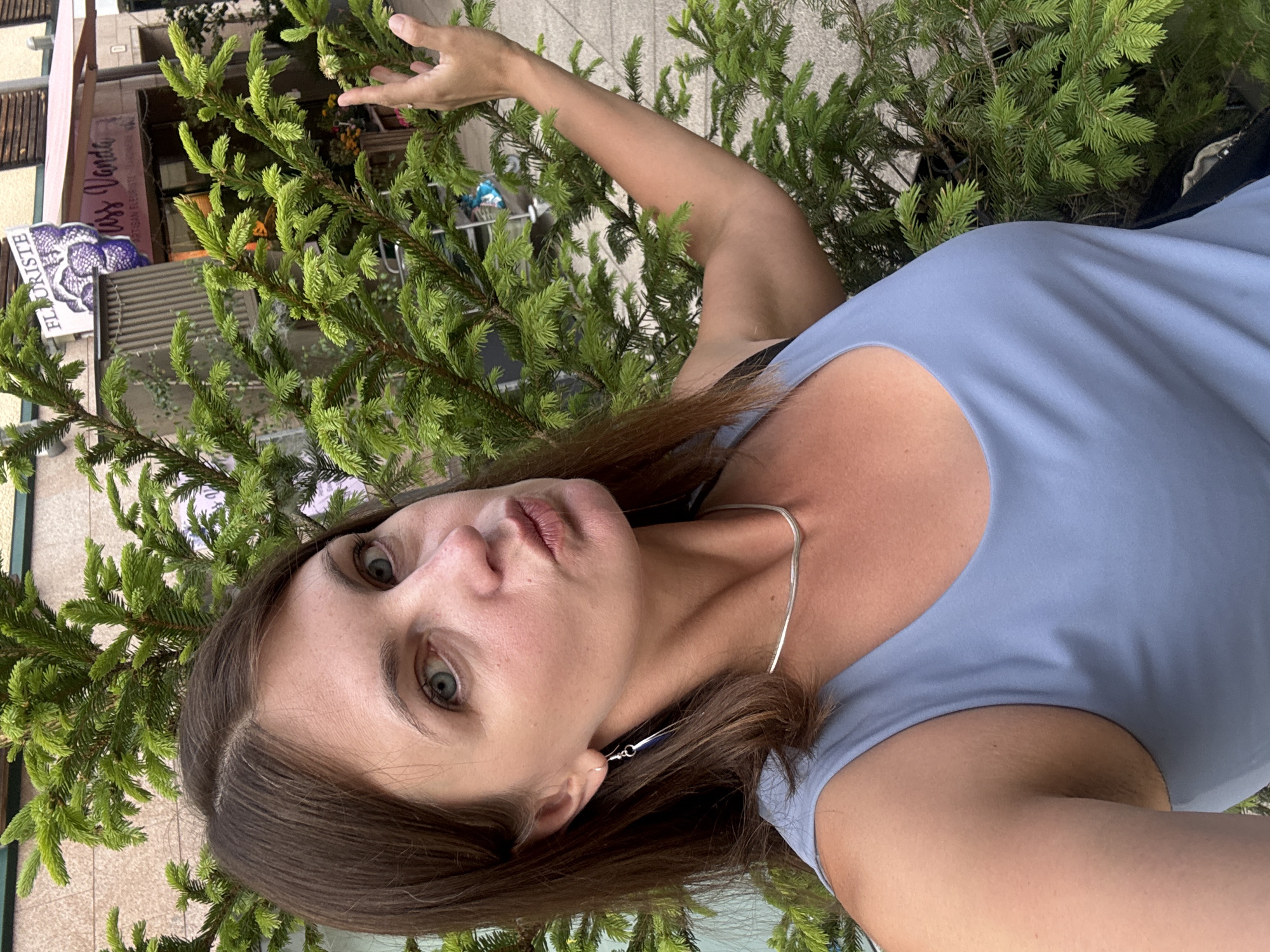 A woman with brown hair, wearing a light blue tank top and a silver necklace, poses for a selfie outdoors. She is positioned in front of a large green coniferous tree, with its branches extending across the frame. In the background, a building with signage for "FLORISTE" and "Miss Vanda Artisan Fleuriste" is visible. The woman is puckering her lips and her right arm is bent, with her hand near the tree's branches. The overall scene suggests an urban environment, possibly a street in Les Rousses, France, given the florist shop.
