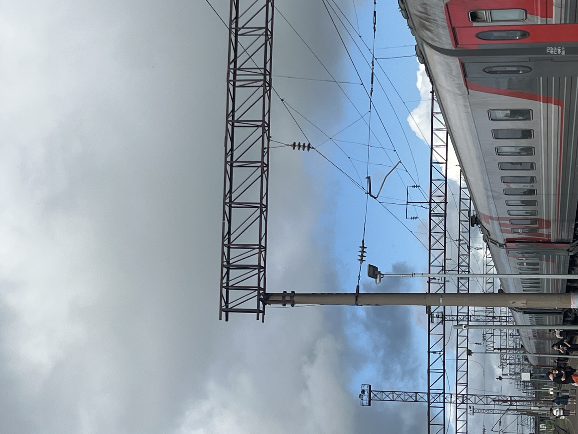 This image depicts a train station in Kanash, Russia, under a cloudy sky. A modern passenger train, primarily silver with red accents, is stopped at the platform. Several individuals are visible on the platform, some standing and some walking. The station infrastructure includes overhead power lines supported by metallic beams and concrete poles, as well as a street lamp. The sky is a mix of grey clouds and patches of blue.