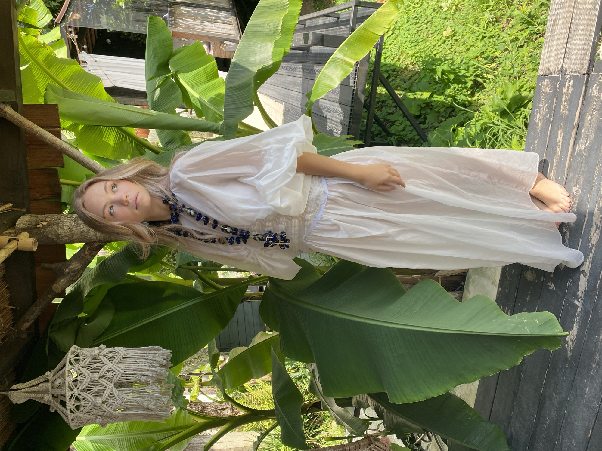 The image depicts a young woman with blonde hair, wearing a sheer white dress and a beaded necklace, standing outdoors amidst lush greenery and banana leaves. Her pose suggests a moment of casual relaxation, possibly a photoshoot. The setting appears to be a tropical or subtropical environment, with abundant foliage and natural light indicating a daytime scene, likely sunny. The wooden deck and bamboo accents hint at an outdoor living space or a resort. The overall mood is serene and natural. There is no visible text in the image.