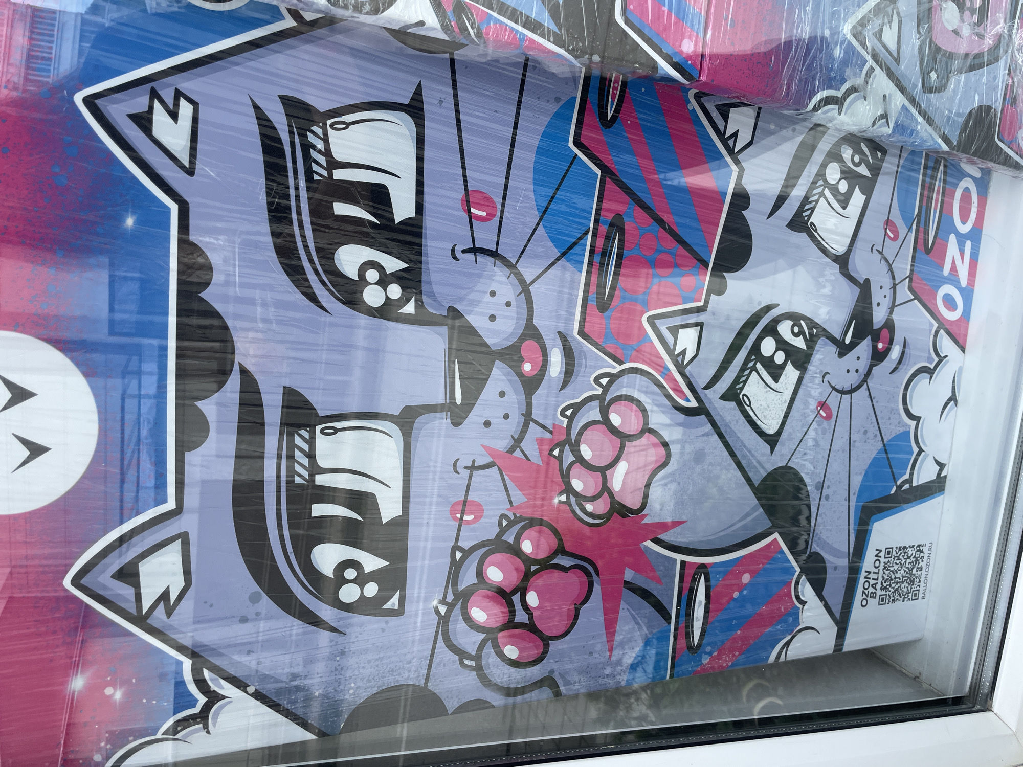 The image depicts a stack of wrapped posters featuring a cartoonish cat design, displayed behind a window in what appears to be Olginka, Russia. The posters showcase a stylized, lavender-grey cat character with bold black outlines and white accents. The background incorporates elements of pink, blue, and white, with geometric shapes like stars, stripes, and polka dots. A QR code is visible on the edge of the topmost poster, labeled with the text "OZON BALLON BALLON.OZON.RU." The posters are positioned close to the glass, and the window frame is white. The reflection on the glass obscures some details.