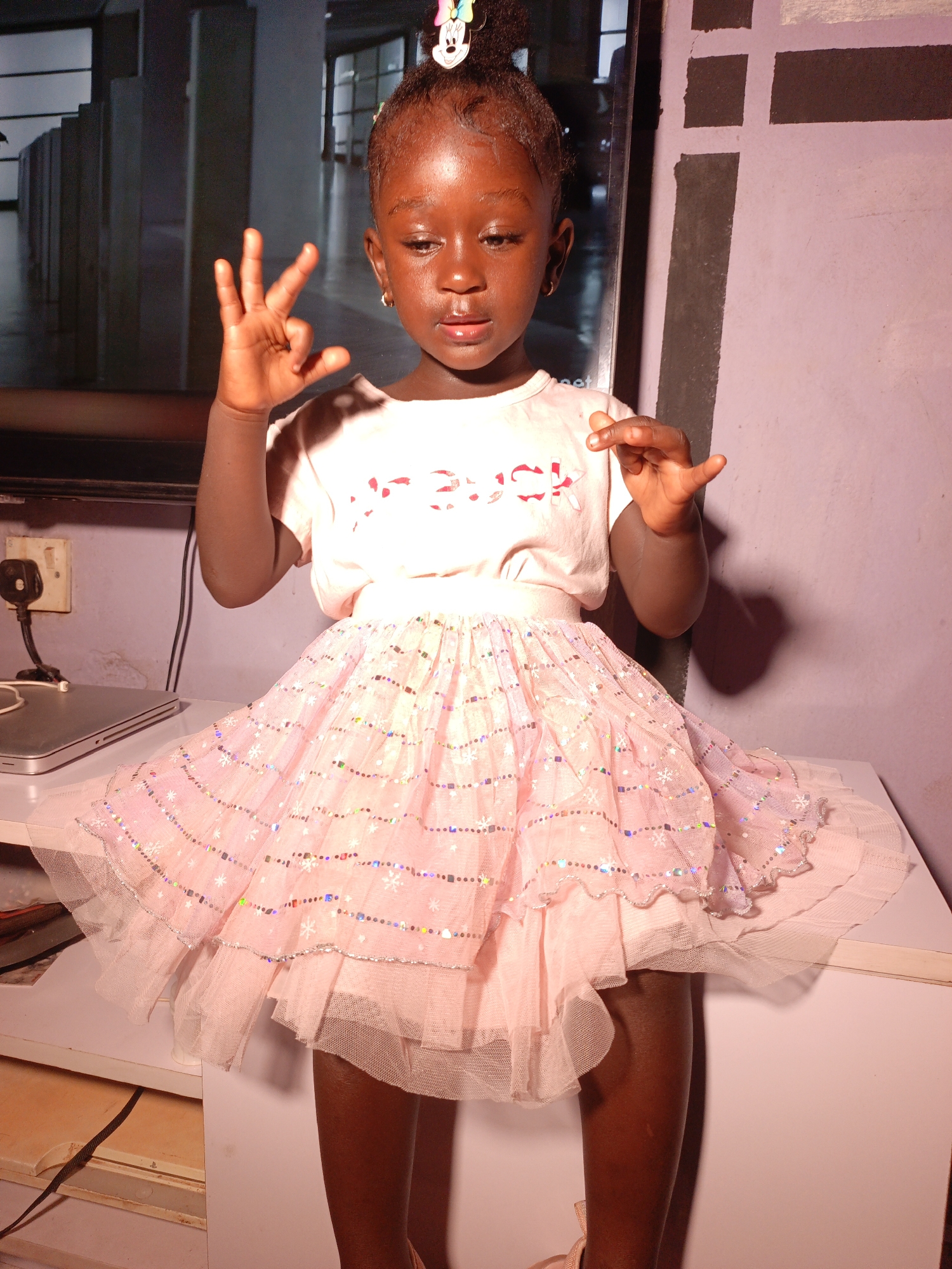 A young, dark-skinned girl stands on a white surface. She wears a light pink t-shirt with faded, red text, and a multi-layered pink tutu skirt adorned with glittery patterns. Her hair is styled in a high bun with a hair accessory. She is holding up her left hand with her index, middle, and ring fingers extended, while her right hand has her index finger extended.
Behind her, there's a dark television screen and a wall with irregular black stripes. To the left of the girl, on the same white surface, is a silver laptop and a power outlet. The location is indoors, the origin location is Akwanga, Nigeria.