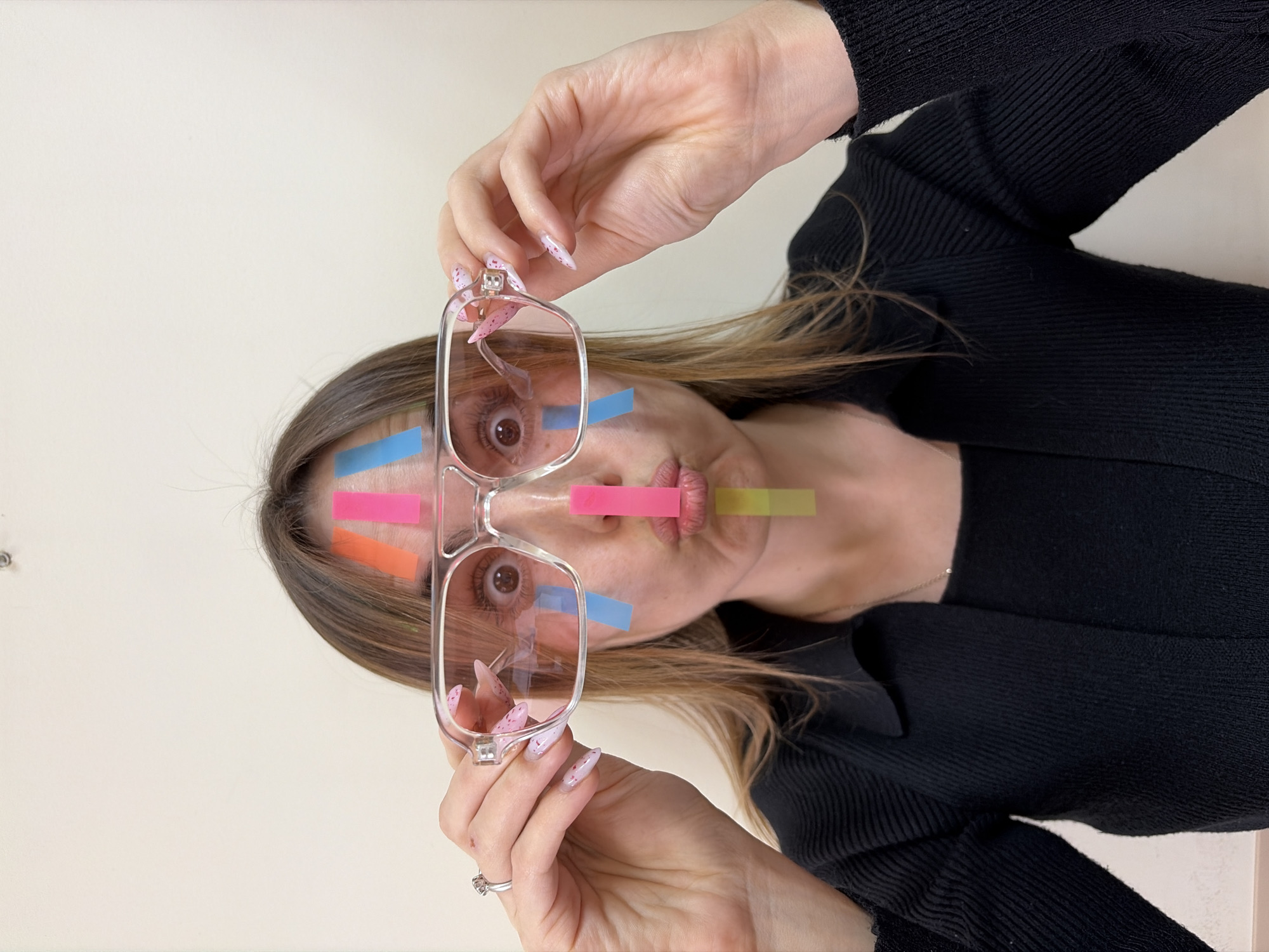 A fair-skinned woman with light brown hair poses holding clear-framed eyeglasses in her right hand. Her face is partially covered with multiple colored adhesive notes: orange, pink, blue and yellow. She is wearing a black, tight-fitting sweater. Her fingernails are long and have a pink and white pattern. A silver ring is visible on her left hand. The background is a plain off-white wall. The image does not provide information on the location of the subject in Izhevsk, Russia.