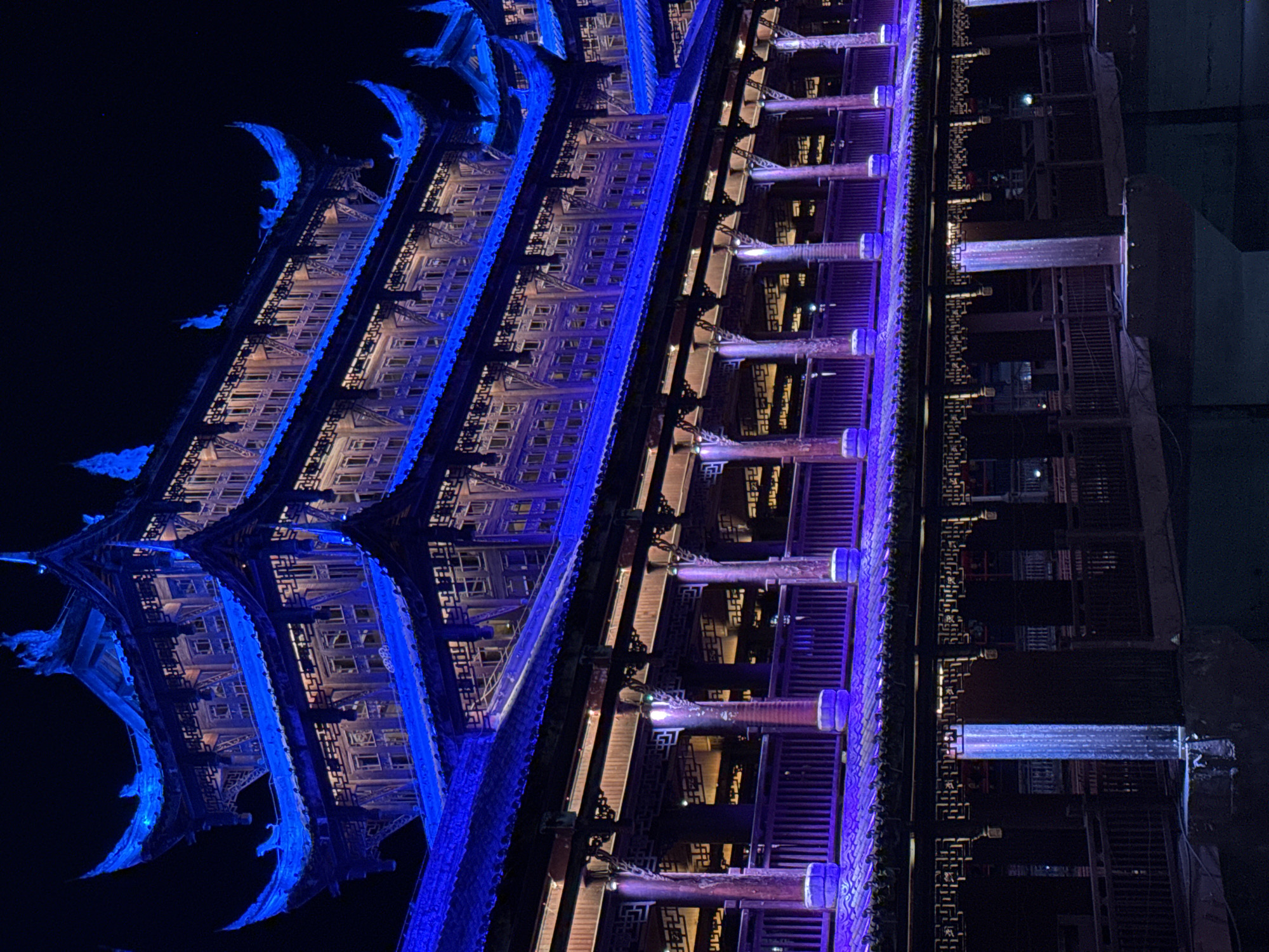 A long-exposure, low-angle shot captures a multi-tiered building at night in Qianjiang, China. The left side of the structure is highlighted in bright blue, contrasting with the right side that is illuminated in varying shades of purple. Architectural details are clearly visible, including the roof's design with its intricate upturned eaves. The building itself appears dark, and the background is a stark black sky, creating a dramatic contrast.