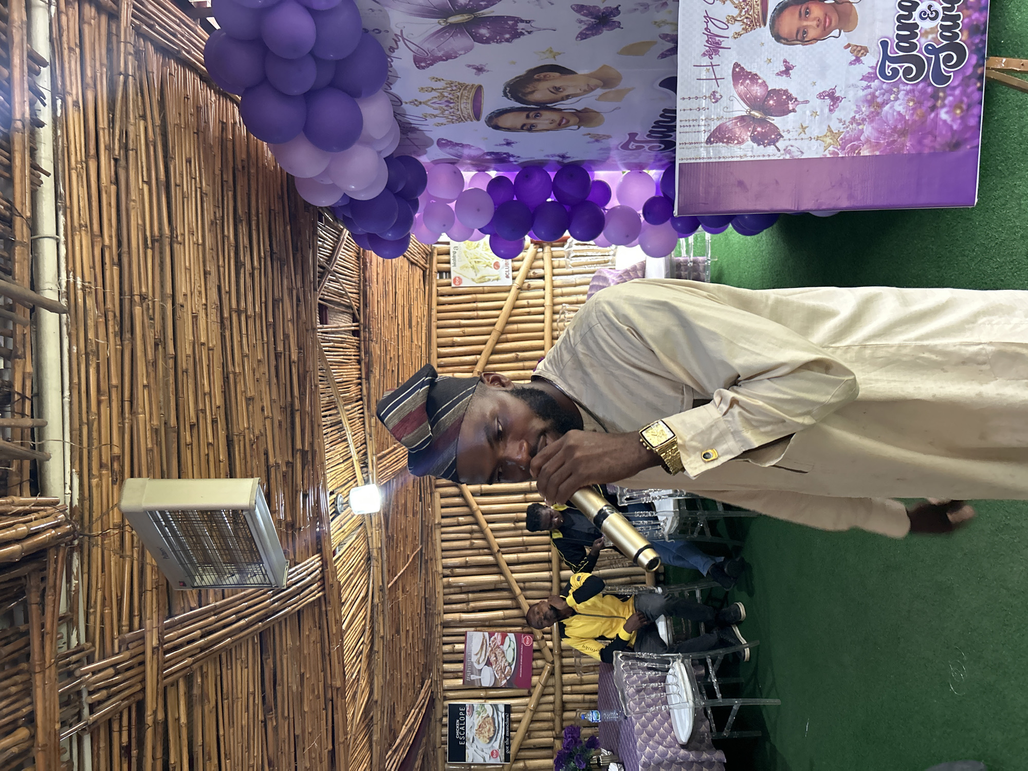 In Bar Jirgi Summa, Nigeria, a man in a cream-colored traditional outfit and a striped cap holds a golden microphone to his lips. He stands on a green carpeted floor in front of a bamboo-paneled wall. To his left, two men are seated at a table covered with a purple patterned tablecloth, adorned with clear plastic chairs. In the background, there are posters depicting food items and event decorations, including purple balloons and banners with images of butterflies and two young girls, with "Tana & Sara" inscribed. A ceiling fan is visible overhead.
