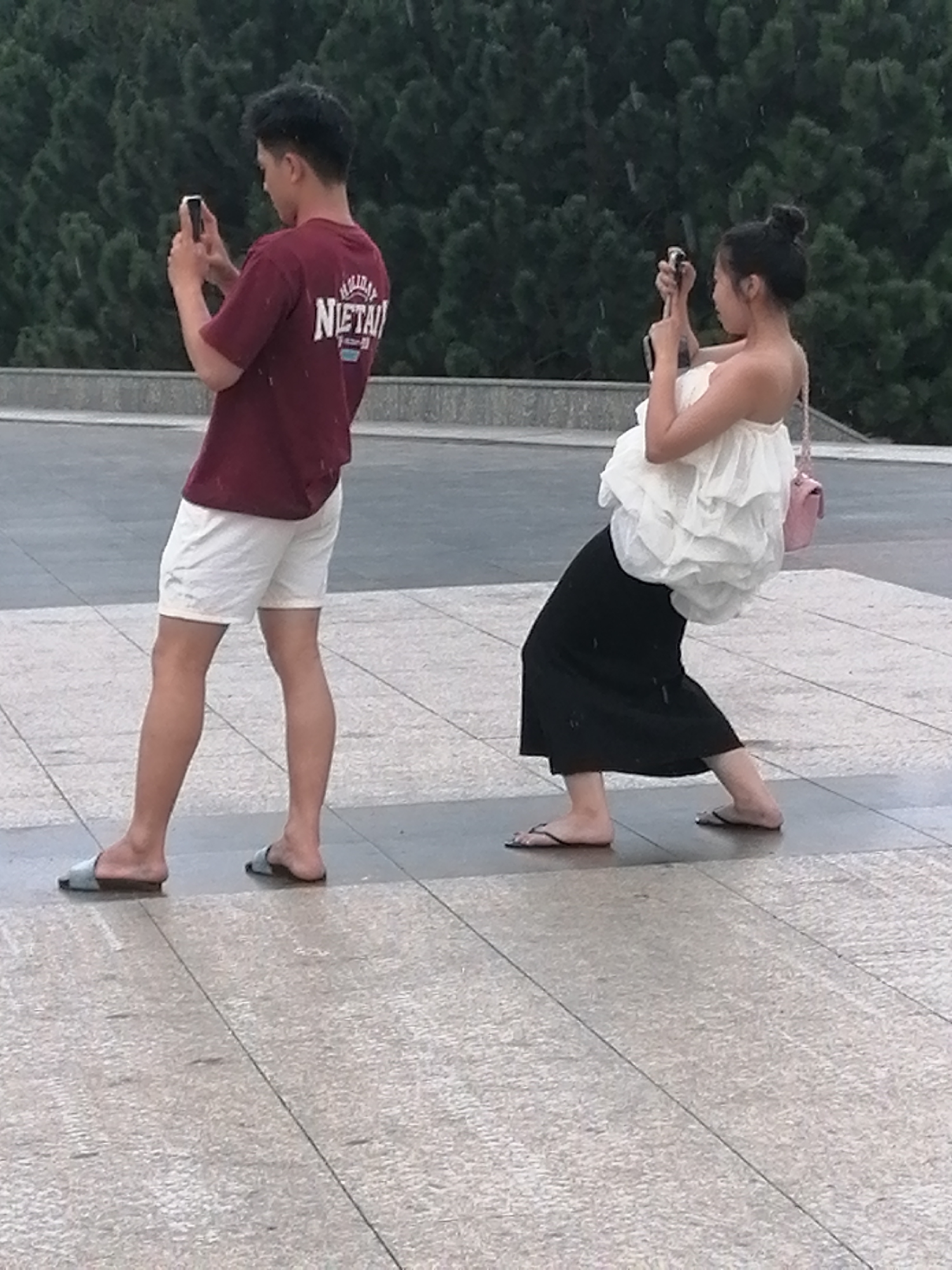 In Da Lat, Vietnam, two individuals are captured in a moment of leisure on a paved public area, possibly a plaza, with a backdrop of lush green trees. The scene unfolds on a bright, overcast day, with the ground exhibiting a slight sheen, suggesting recent moisture or a damp surface.
The individual on the left, a young male, stands facing left, engrossed in his smartphone. He is dressed in a maroon t-shirt with white lettering and crisp white shorts, paired with light blue slide sandals. His stance is relaxed, with one leg slightly forward.
To his right, a young female is depicted in a dynamic pose, leaning back and looking towards her own smartphone, which she holds in both hands. She wears a white, strapless ruffled top and a flowing black skirt, complemented by black flip-flops. A small pink handbag is slung over her right shoulder. Her posture suggests an engagement with the digital world, perhaps capturing a photo or video.
The ground beneath them is a mosaic of square and rectangular paving stones, a common feature in urban Vietnamese settings. The overall impression is one of everyday life, where technology and personal leisure intersect within a serene, natural environment characteristic of Da Lat.