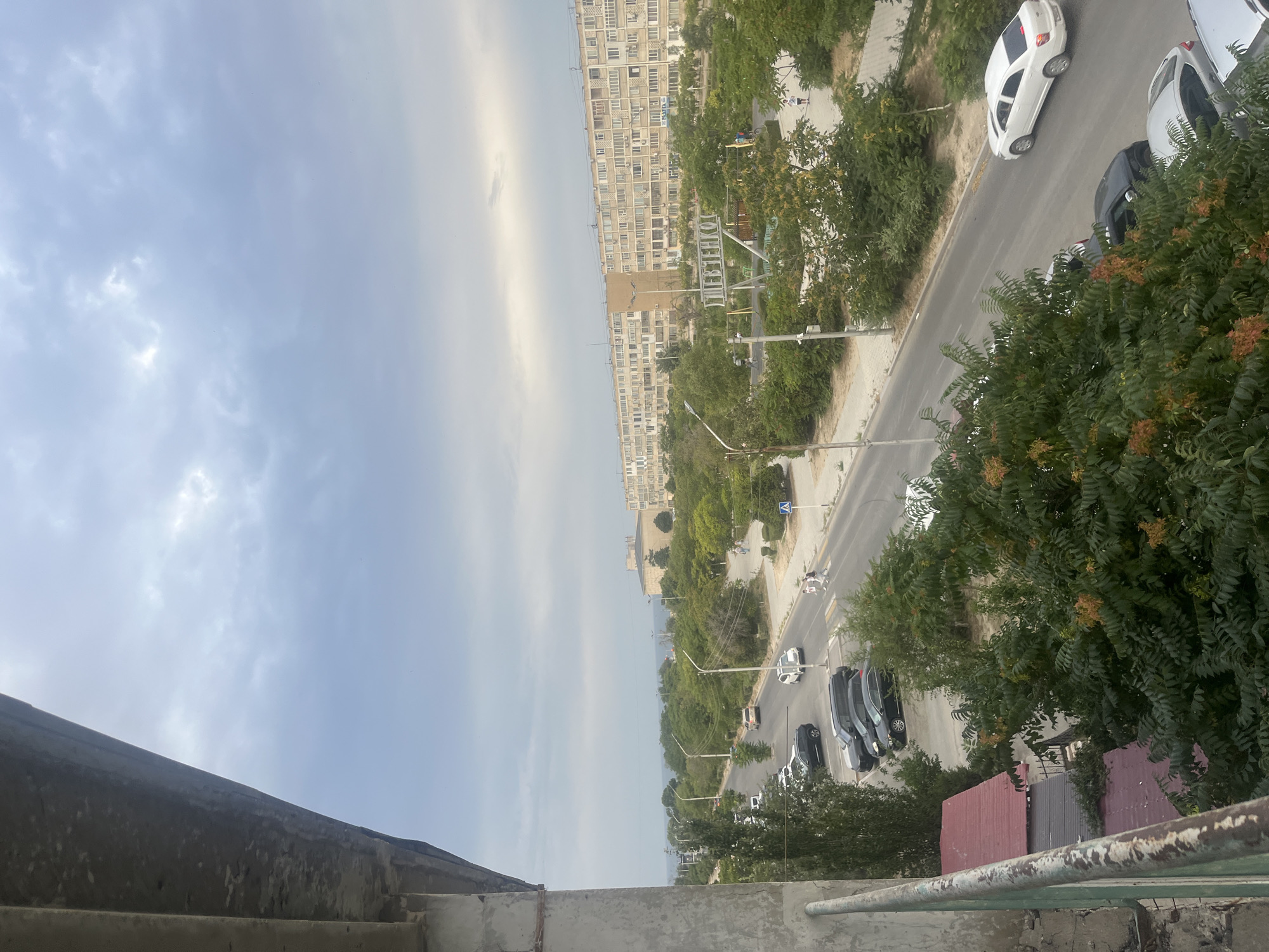 The image shows a street scene in Aktau, Kazakhstan. A long, multi-story apartment building dominates the background, with numerous windows and balconies. A wide street runs through the middle ground, with cars parked on the side and driving on the road. A park area with trees, playground equipment, and a sign with Cyrillic text is visible. In the foreground, a balcony railing and some dense foliage are prominent. The sky is overcast with grey clouds. Several people are visible walking along the sidewalk and crossing the street. The overall scene suggests a residential urban environment.