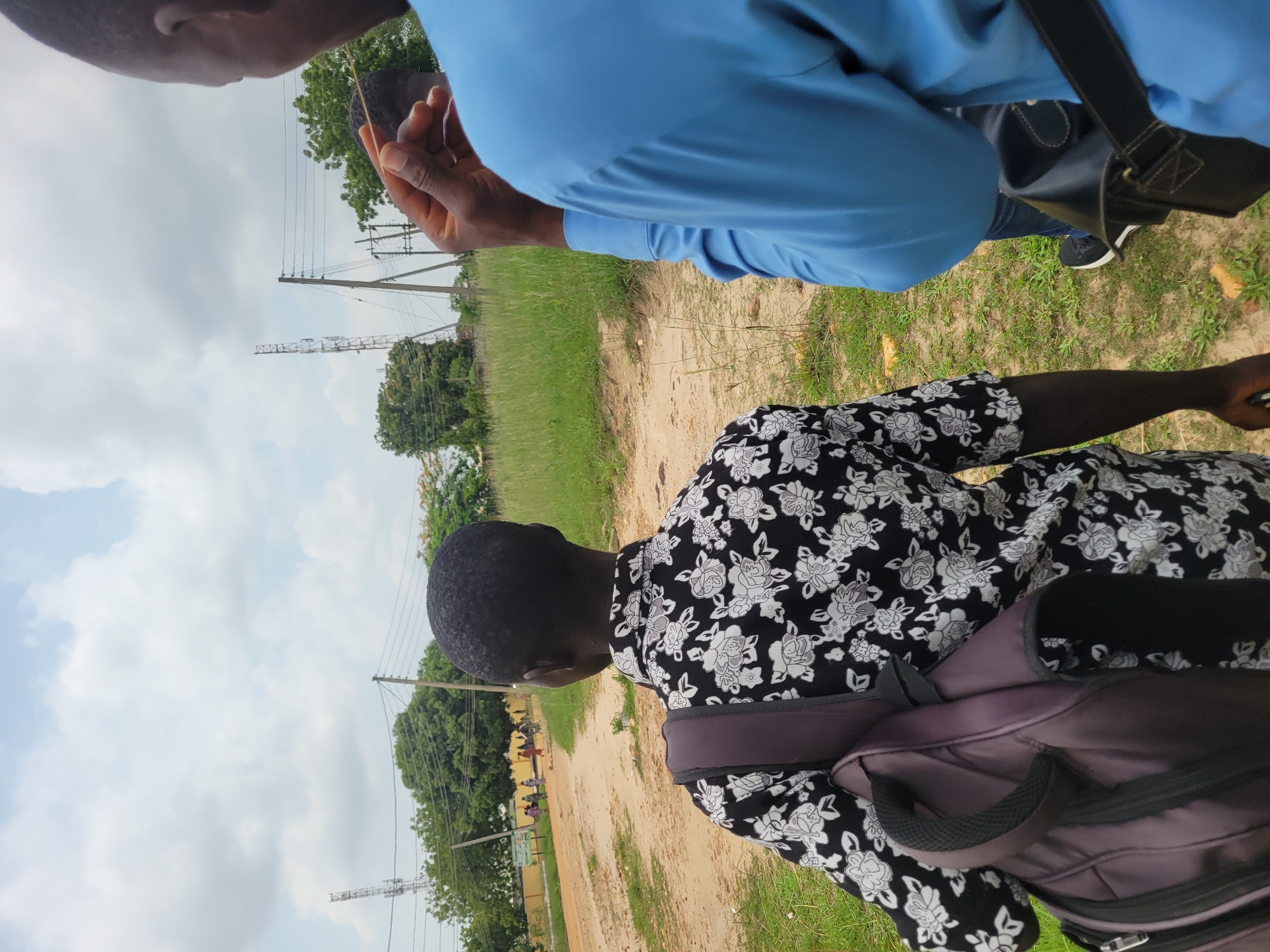 Two individuals are positioned in the foreground of a dirt road, which curves towards the background of the image. The person on the left is viewed from behind, wearing a black and white floral-patterned shirt and a dark purple backpack. Their head is turned slightly to the right. The person on the right, whose back is to the viewer, is wearing a blue shirt and is holding a thin, light-colored object in their right hand. The environment consists of dry, brown grass on the right side of the road and a patch of taller, greener grass on the left. In the background, trees with lush green foliage are visible. Several telecommunication towers stand tall against a cloudy sky. A group of people are walking on the dirt road in the distance, and a building with a yellow exterior and a green sign can be seen to the left of the road. The scene appears to be outdoors in a semi-rural area of Keffi, Nigeria.