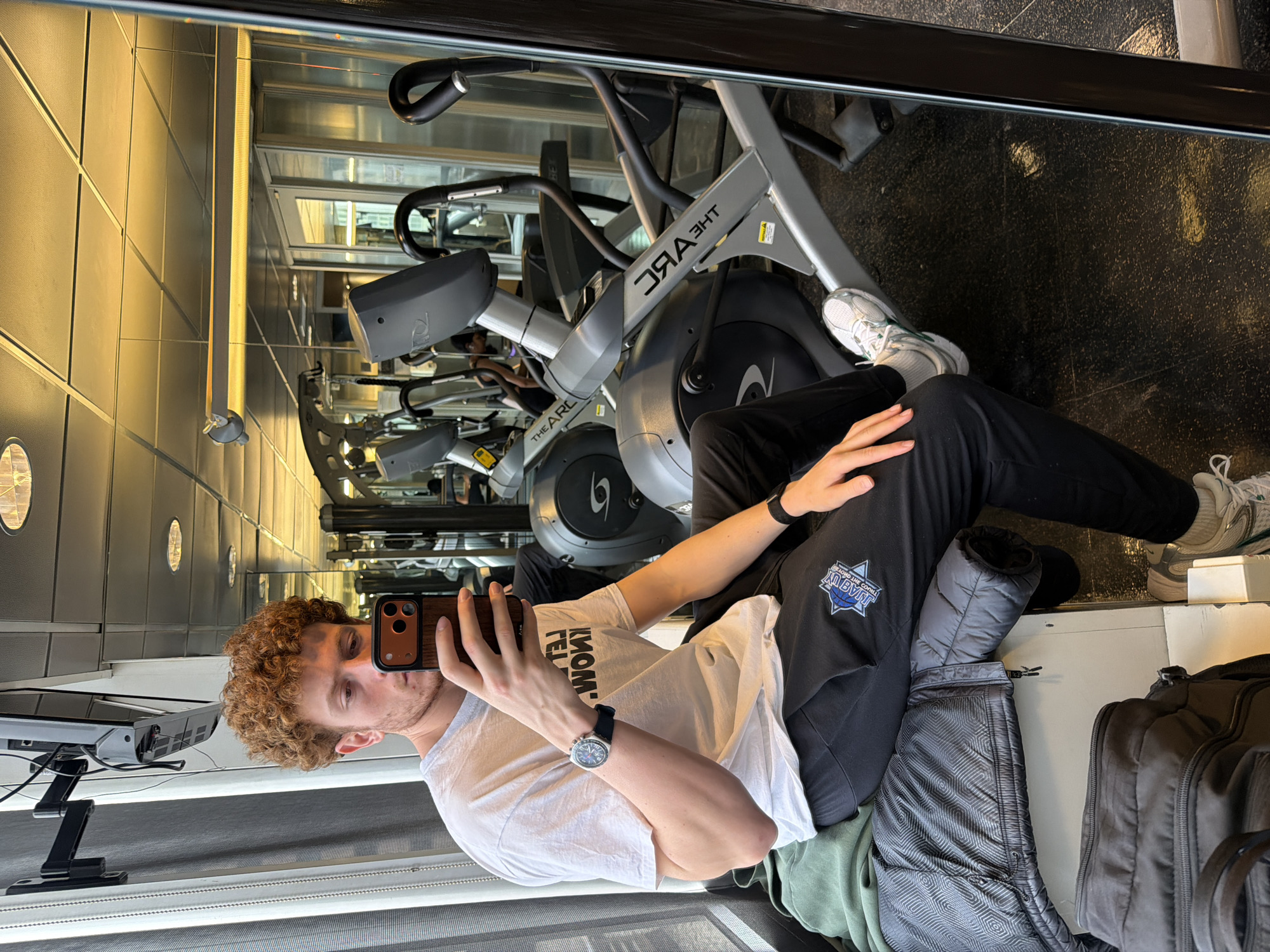 A selfie-style photograph captures a man with curly red hair in a gym setting. The man is holding a smartphone with a brown case and is positioned towards the left side of the image. He is wearing a white t-shirt, black pants with a logo, and a wristwatch. His clothing is layered with additional garments and a dark-colored bag nearby. The gym is equipped with exercise machines, including a visible "The ARC" elliptical trainer. A mirror reflects the gym's interior, showcasing rows of equipment and other individuals exercising. The environment suggests a modern fitness center setting, possibly located in New York City, United States of America.
