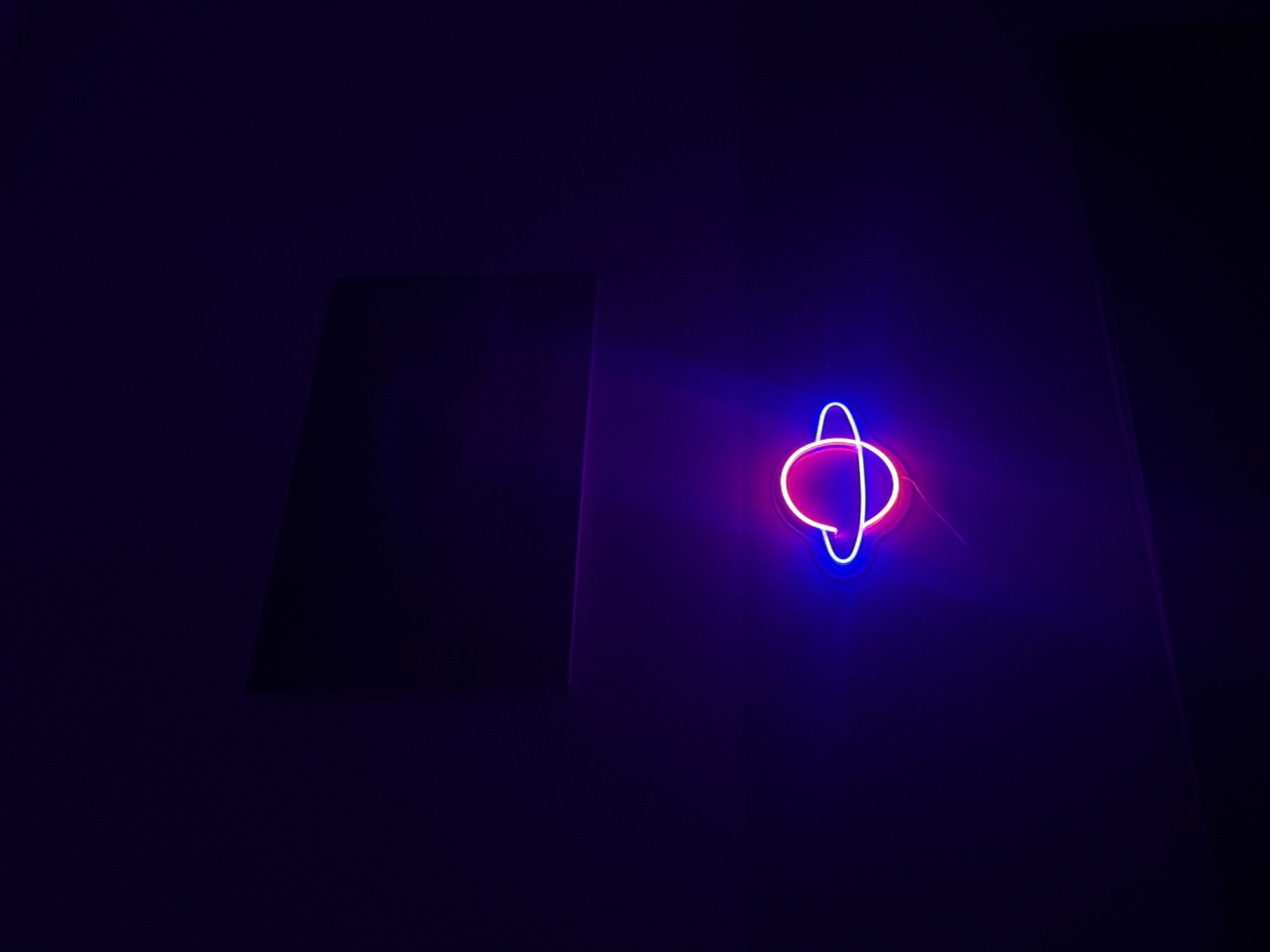 In a dimly lit room, a neon sign shaped like a planet with rings glows. The planet is rendered in magenta, and the rings are in bright blue. Above the planet, a dark rectangular object hangs on the wall, its surface reflecting a faint purple light. The room is painted a deep purple, and the overall ambiance is dark and atmospheric. The image was taken in Nha Trang, Vietnam.