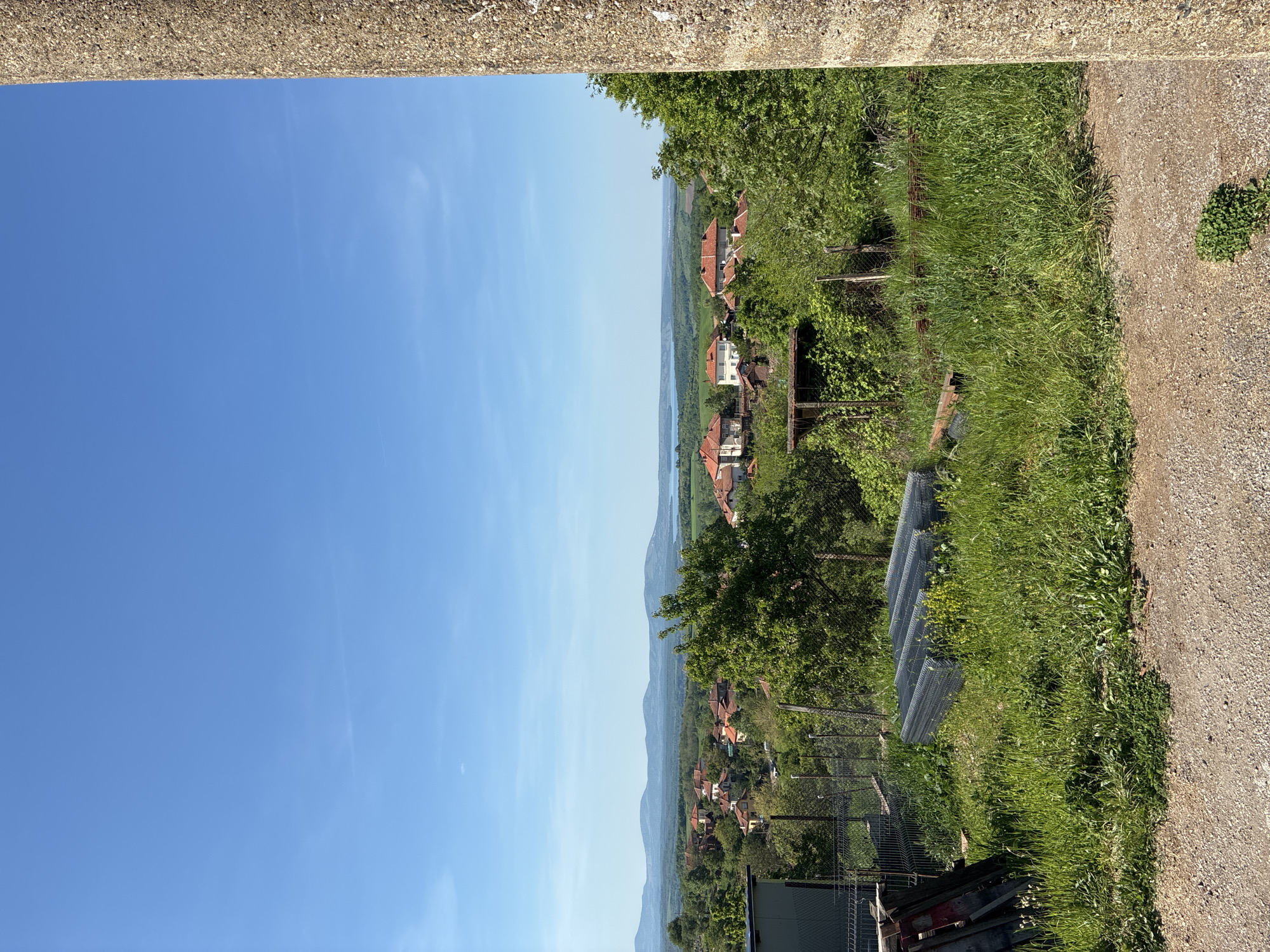 The image shows a view in Bokilovtsi, Bulgaria. The sky is clear and blue. The foreground features a concrete structure and overgrown grass. In the distance, there are houses with red roofs amidst green vegetation, suggesting a village setting. Hills or mountains are visible in the far background. Wire mesh and wooden planks lay in the grass.