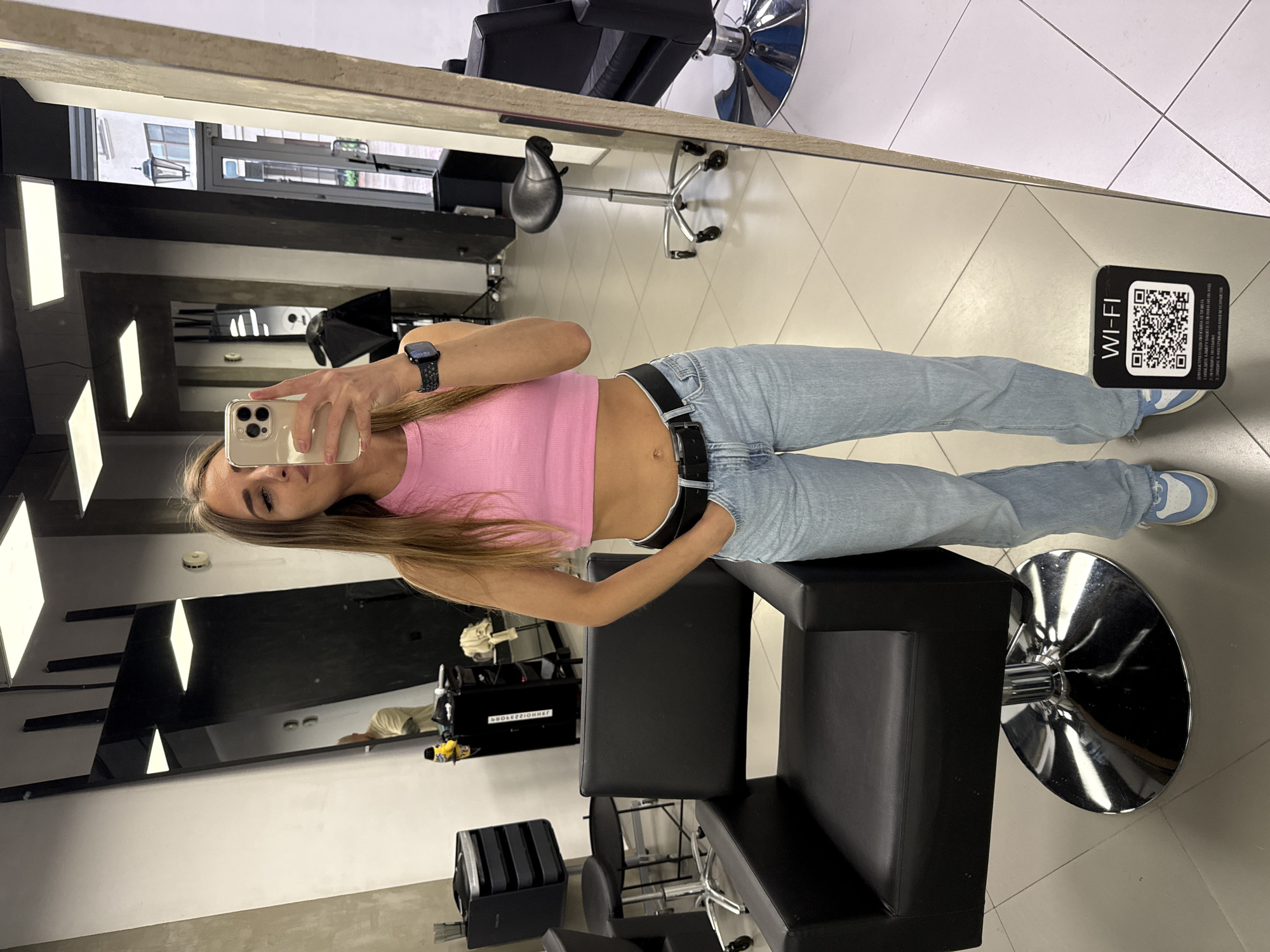 The image is a mirror selfie taken in what appears to be a hair salon. The person in the photo is a young woman with long brown hair, wearing a pink crop top, light blue jeans, and a black belt. She is holding a smartphone in her left hand, presumably to take the picture. A black smartwatch is visible on her left wrist.
Behind her are various salon chairs, mirrors, and equipment, suggesting a professional hairstyling environment. The decor is modern, with black and gray color schemes dominating the interior design. A QR code with the label "WI-FI" is visible on the upper right side of the mirror.
Based on the architectural details visible in the mirror's reflection, the location could be in Saint Petersburg, Russia. The reflection reveals a building with a distinct architectural style common in the city.