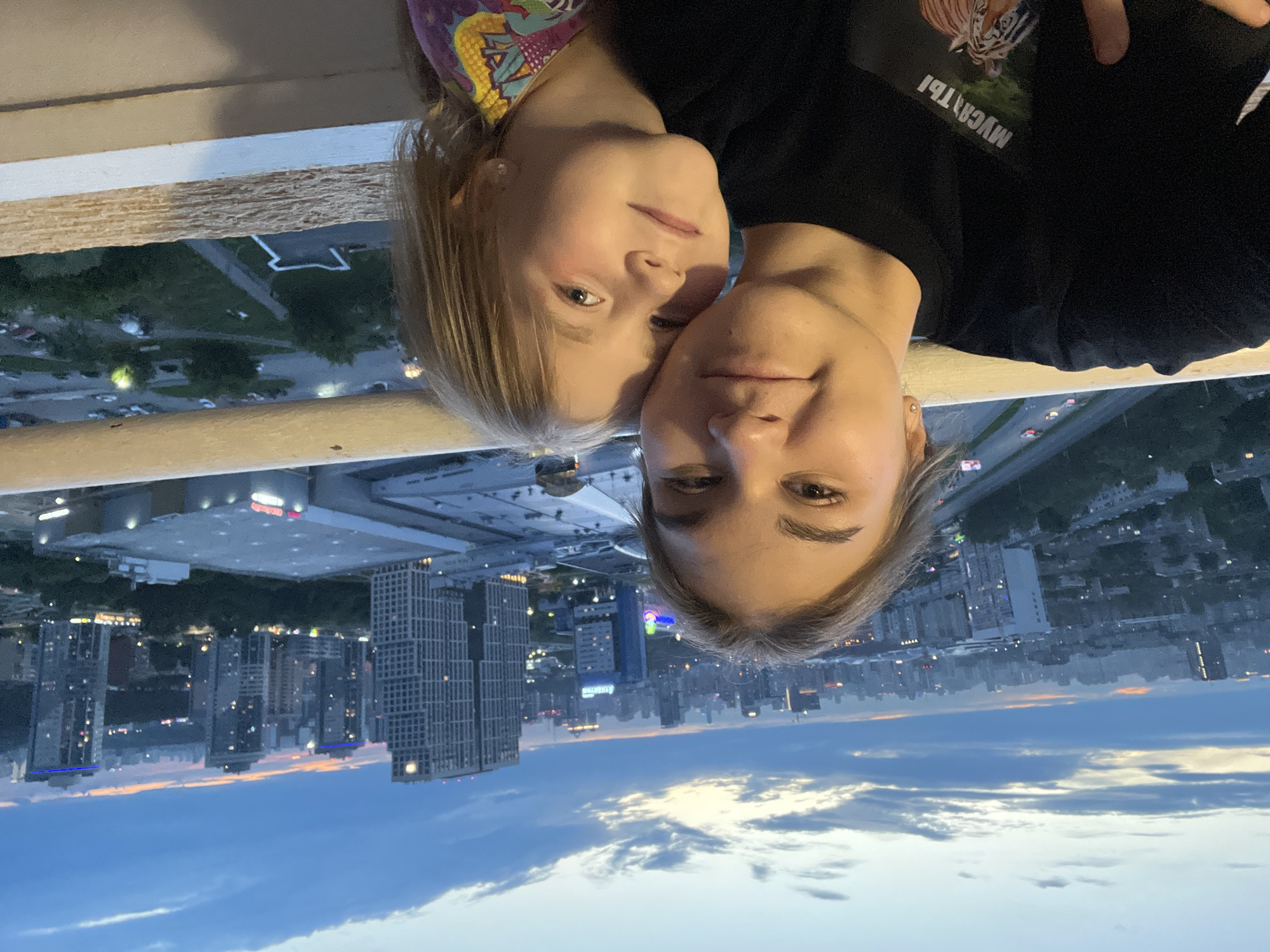 In this selfie taken at dusk, two females are positioned in the foreground against a backdrop of a sprawling city skyline. The older female, on the left, has her arm around the younger female on the right, who is leaning into her. Both are looking towards the camera. The younger female has blonde hair and is wearing a multi-colored shirt. The older female is wearing a black t-shirt with a graphic print on the front. The cityscape features numerous high-rise buildings and a road with cars visible in the mid-ground. The sky is cloudy with hints of orange and pink from the setting sun. The image appears to have been taken from a high vantage point, likely a balcony or rooftop, overlooking the city of Yekaterinburg, Russia.