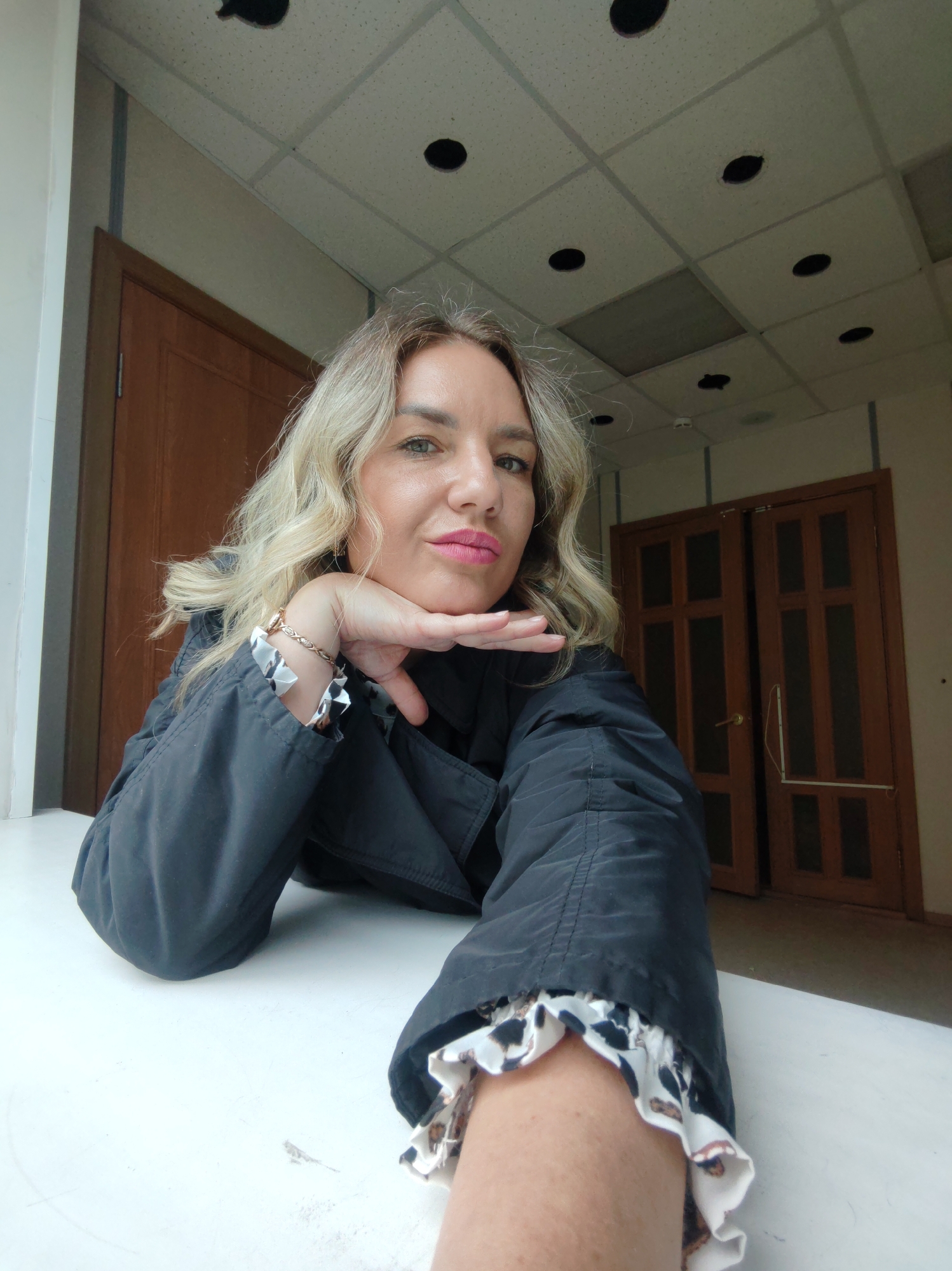 A woman with blond wavy hair and pink lipstick is resting her face on her hand while taking a selfie. She is wearing a black jacket with a ruffled sleeve. Behind her are two sets of brown doors and a suspended ceiling with circular holes. The location is likely indoors, but specific location information is unavailable.