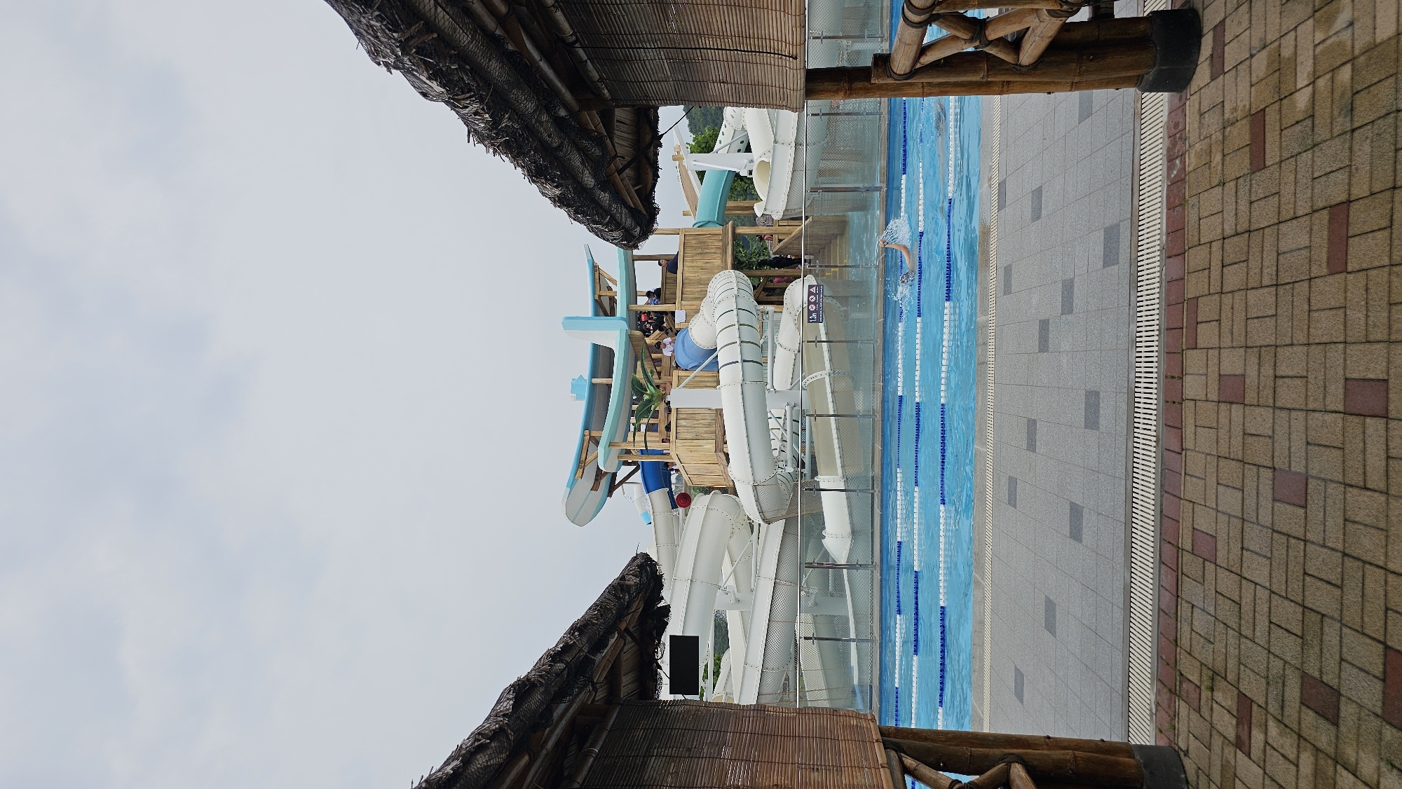 The image depicts an outdoor water park on a cloudy day. In the foreground, two thatched-roof structures with bamboo walls frame the view. Between them, a swimming pool with lane markers is visible, and a person is swimming. Beyond the pool, a large water slide complex with white and blue slides is prominently featured. The complex is made of wood and has multiple levels. Some people are visible on the platforms of the water slide structure. The ground in front of the pool is paved with square gray tiles and patterned brickwork. The overall environment suggests a recreational facility. The location is identified as Asan-si, South Korea.