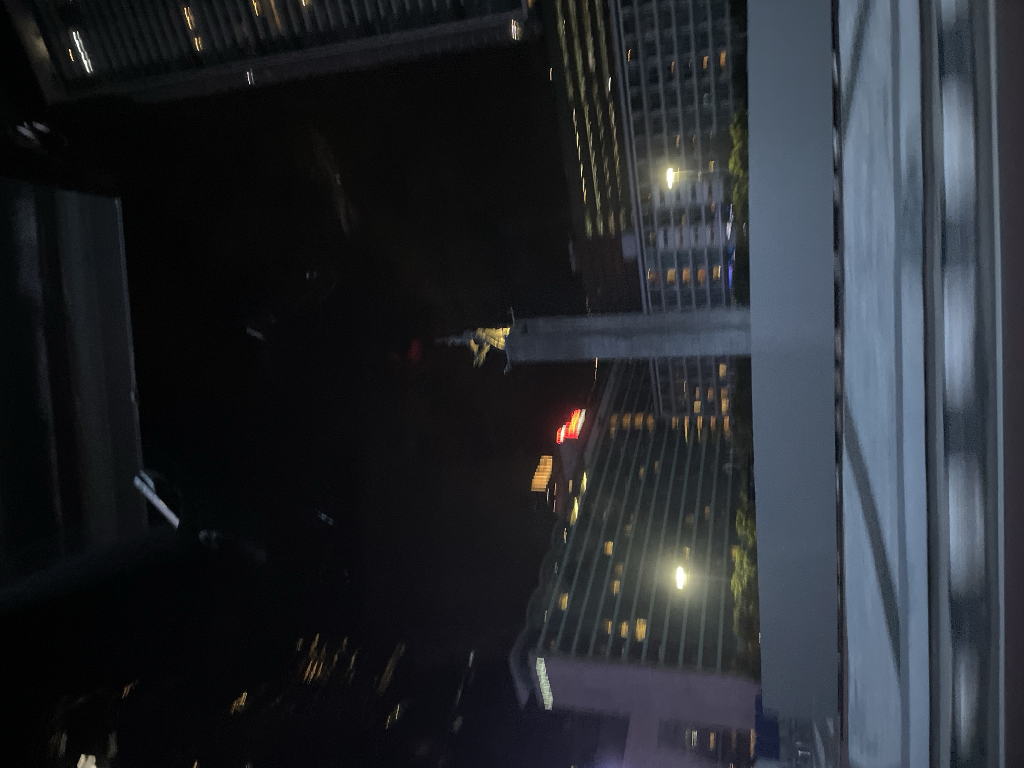 The image shows a night scene in Central Jakarta, Indonesia, as viewed from a high vantage point, likely a balcony or window. Skyscrapers with illuminated windows dominate the background. One building features a red logo at the top. The foreground is dark, with some visible structures, including part of a balcony railing. The image is taken at night.