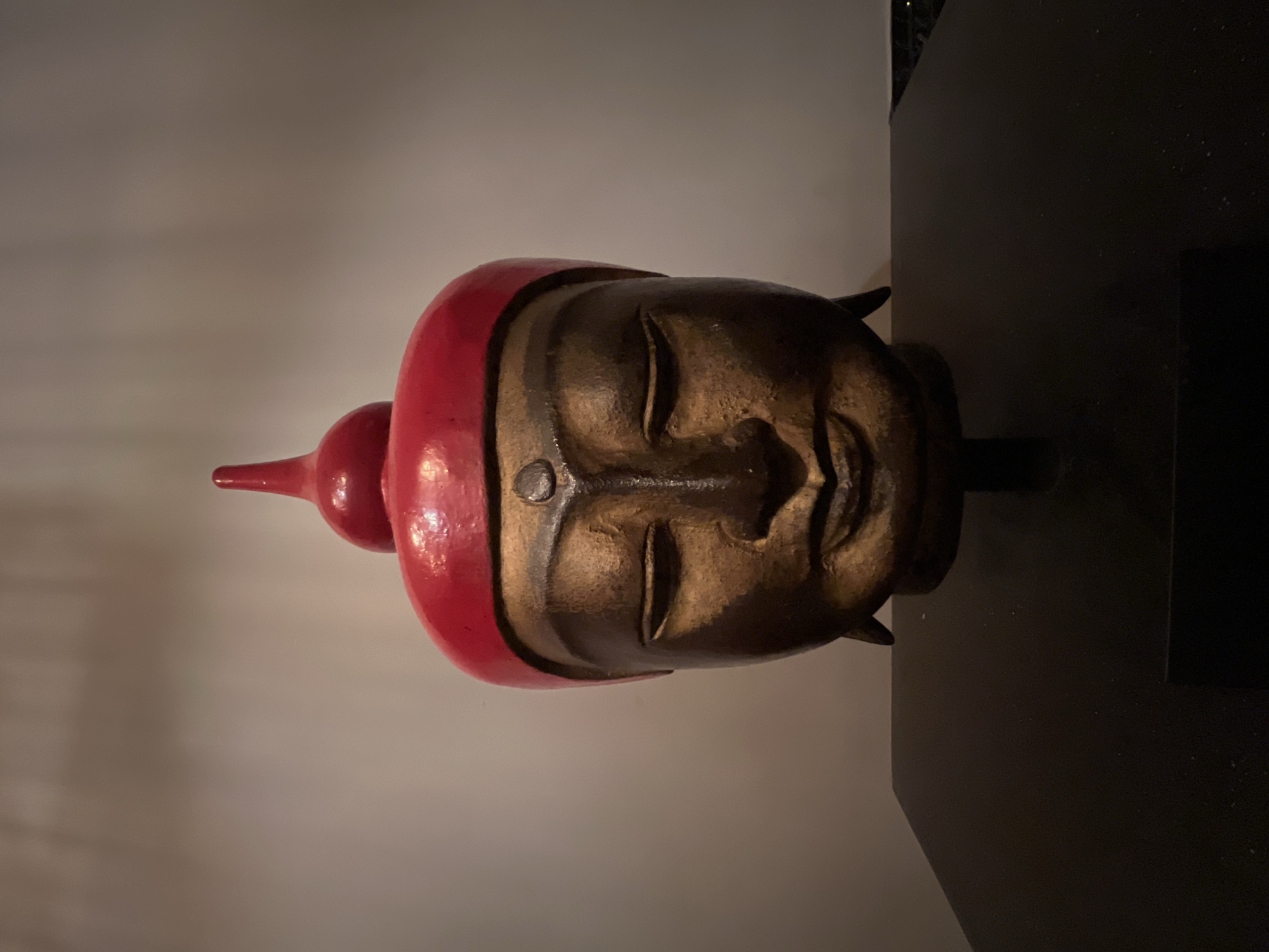 A close-up, head-on, eye-level, medium shot captures a Buddha head sculpture. The sculpture is made of dark wood with a reddish-brown hue, showing visible texture. It is adorned with a bright red headdress that tapers to a point at the top. The Buddha's eyes are closed, and it has a serene, slight smile. A small, dark circular marking is present on its forehead. The sculpture is mounted on a black stand, which rests on a dark, rectangular surface. The background is a plain, light-colored wall. The lighting is dim and focused on the sculpture, creating a shadow effect. The location is not specified in the image or its metadata.
