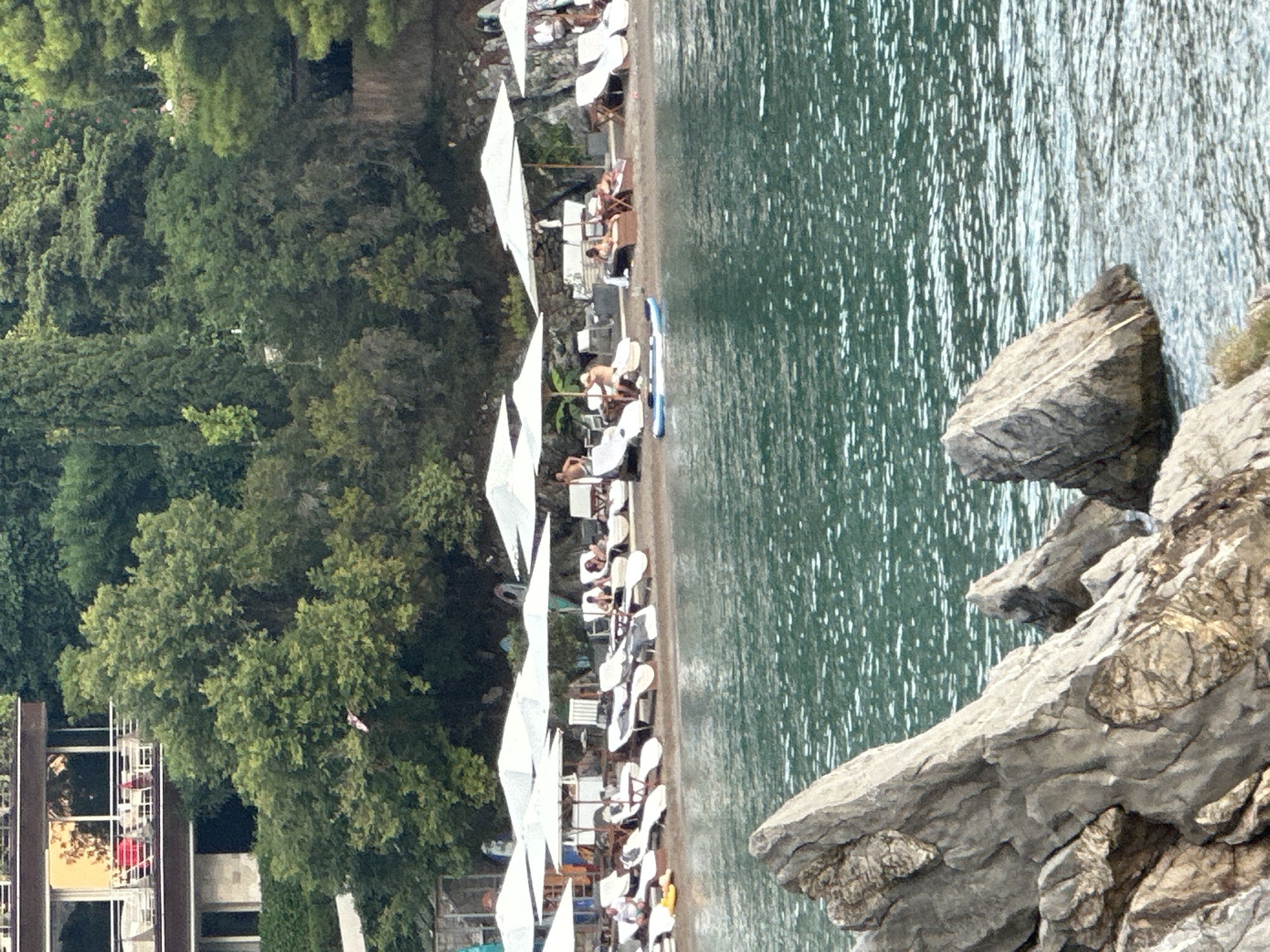 The image depicts a serene beach scene in Budva, Montenegro. In the foreground, weathered rocks emerge from the turquoise waters of the Adriatic Sea, creating a naturalistic contrast to the manicured beach beyond. A shoreline lined with white lounge chairs and umbrellas stretches across the mid-ground, offering shaded respite to beachgoers. Several individuals are seen relaxing on the loungers, some appearing to be sunbathing or engaged in casual conversation. A paddleboard is visible on the sand, suggesting recreational activities. Behind the beach, a lush green hillside is densely forested with trees, and a building with balconies overlooks the scene, hinting at nearby accommodation. The overall atmosphere is one of tranquil leisure, with the natural beauty of the Montenegrin coast providing a picturesque backdrop.