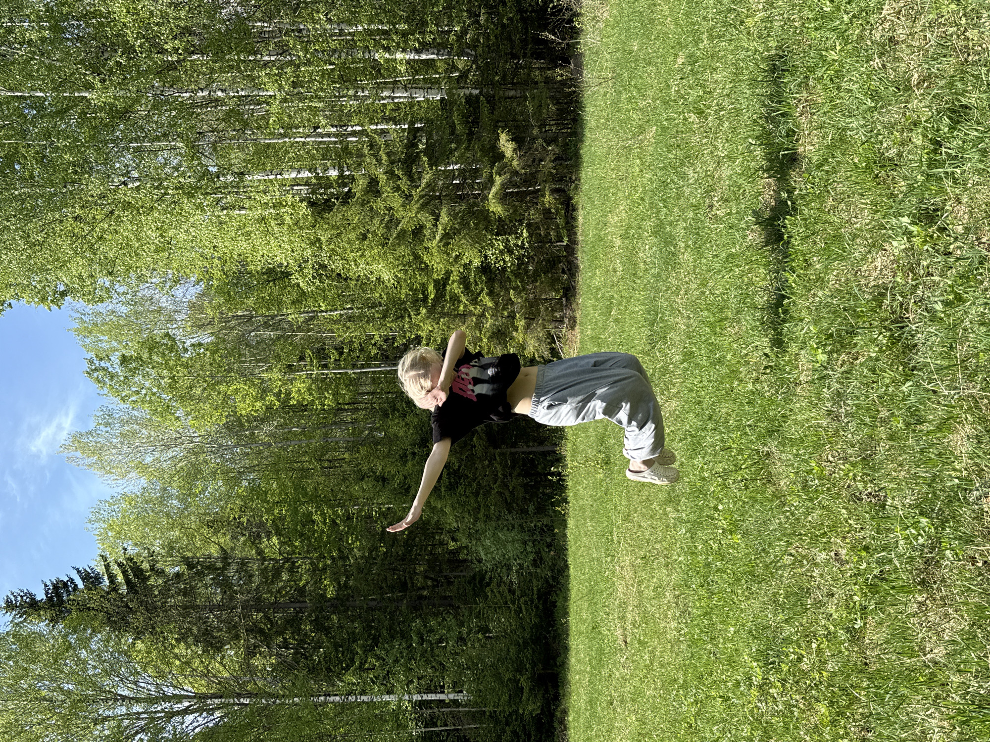 A horizontal, outdoor, full-body shot of a woman on a grassy area adjacent to a forest, with a light blue sky visible in the upper left corner. The scene is in Zhigalki, Russia.
The woman, who appears to be young, is blonde and wearing a black t-shirt, light blue pants, and beige slip-on shoes. She is in motion, with her body bent at the waist. Her right arm is extended downward, and her left arm is bent with her hand near her head.
The grassy area is green and covers the majority of the lower half of the image. A dense forest with birch and evergreen trees forms the backdrop. The lighting is bright and sunny.