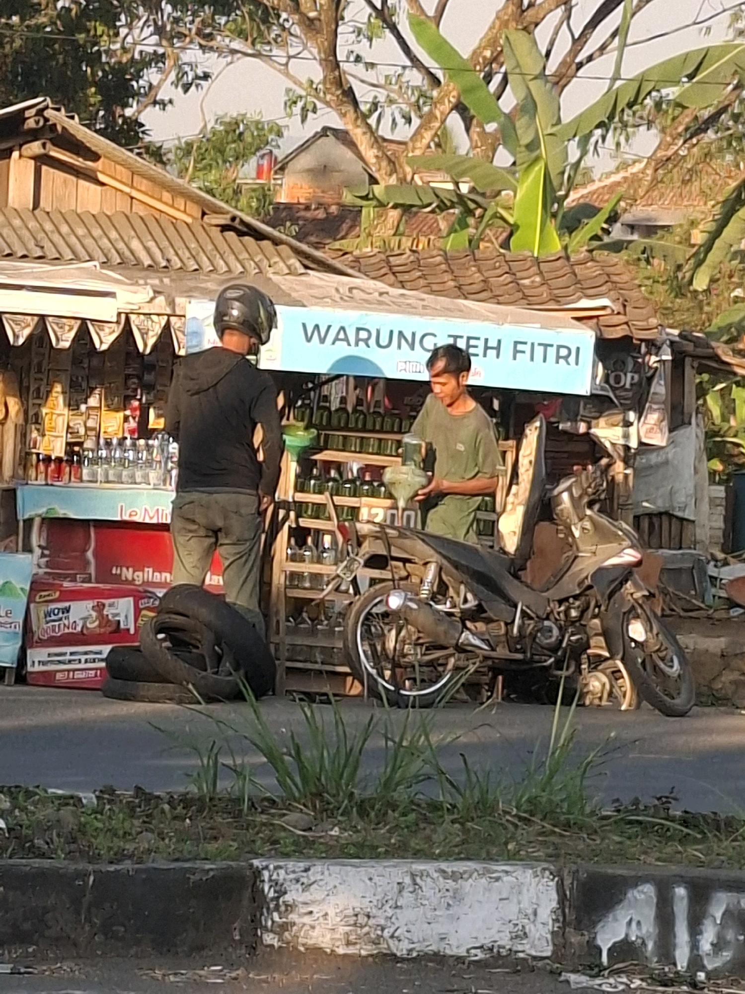 In Cikaret, Indonesia, a small roadside stall, identified by a blue sign reading "WARUNG TEH FITRI," is depicted. The stall appears to sell various bottled beverages. A man wearing a black hooded jacket and dark pants, with a black helmet on his head, stands facing the stall. Another man, wearing a green t-shirt and green pants, is behind the counter, holding a green object. A motorcycle is parked in front of the stall, and stacked tires are visible near the entrance. The scene is set against a backdrop of trees and foliage, with a street in the foreground.