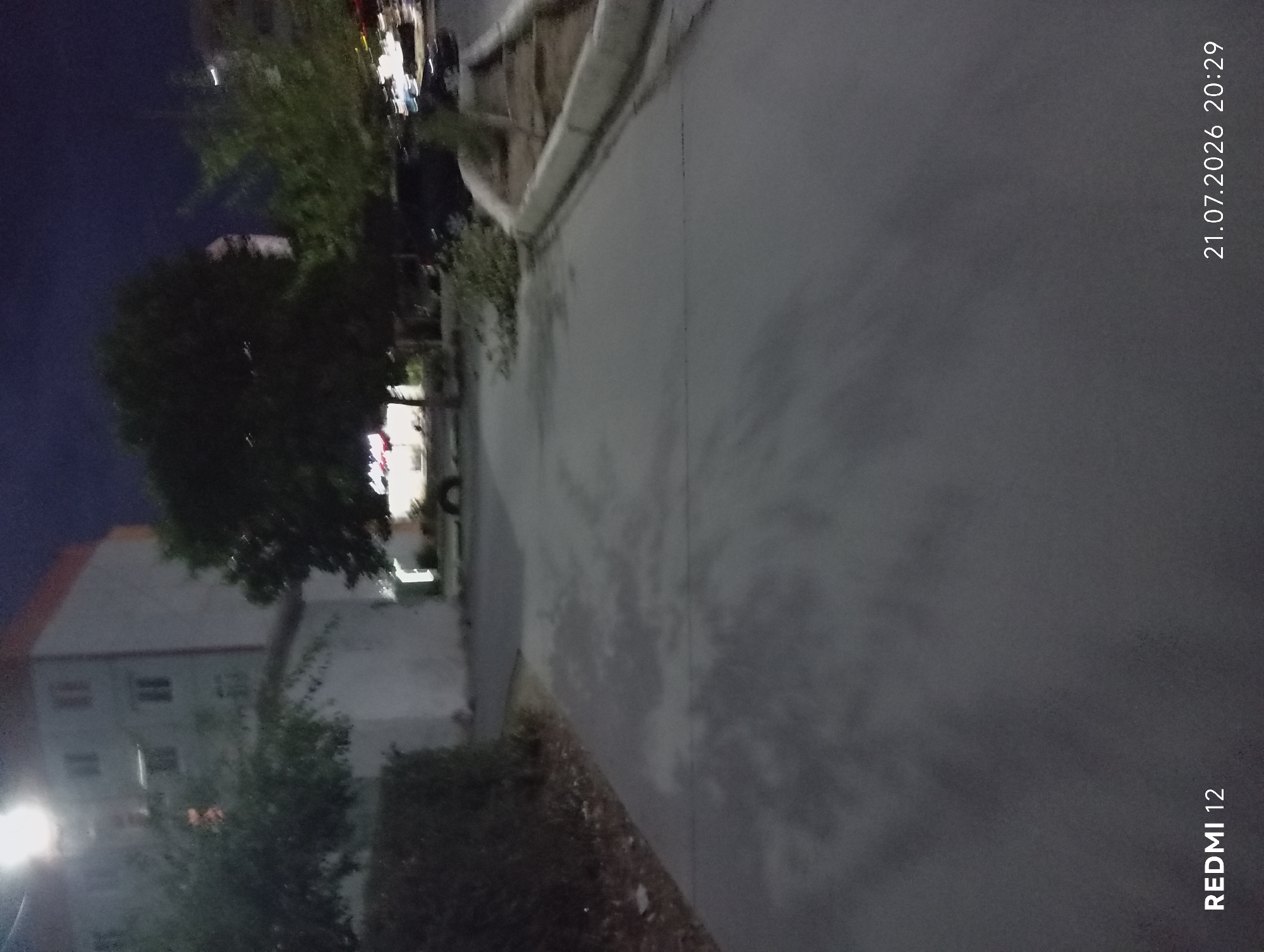 This is a vertically oriented photograph taken at night, likely in Bekabad, Uzbekistan, as indicated by the time stamp. The scene depicts an outdoor environment with a paved walkway or road in the foreground and a building with several windows on the left. A tree with dark foliage is visible in the middle ground, casting shadows on the paved surface. In the background, there appears to be more greenery and some indistinct structures, possibly part of the building or other adjacent structures.
The time stamp "21.07.2026 20:29" suggests it is evening on July 21, 2026. The low light conditions and the presence of shadows indicate it is nighttime. The paved surface appears slightly damp, possibly due to recent rain or dew. A bright light source is visible in the lower left corner, possibly a street lamp or a light from the building.
There are no clearly discernible people in the image. The objects present include the building, trees, and the paved surface. The overall mood of the image is somewhat subdued due to the low light and the nighttime setting.
Visible text includes the date and time stamp "21.07.2026 20:29" and the text "REDMI 12" vertically oriented on the right side of the image, indicating the device used to capture the photograph.