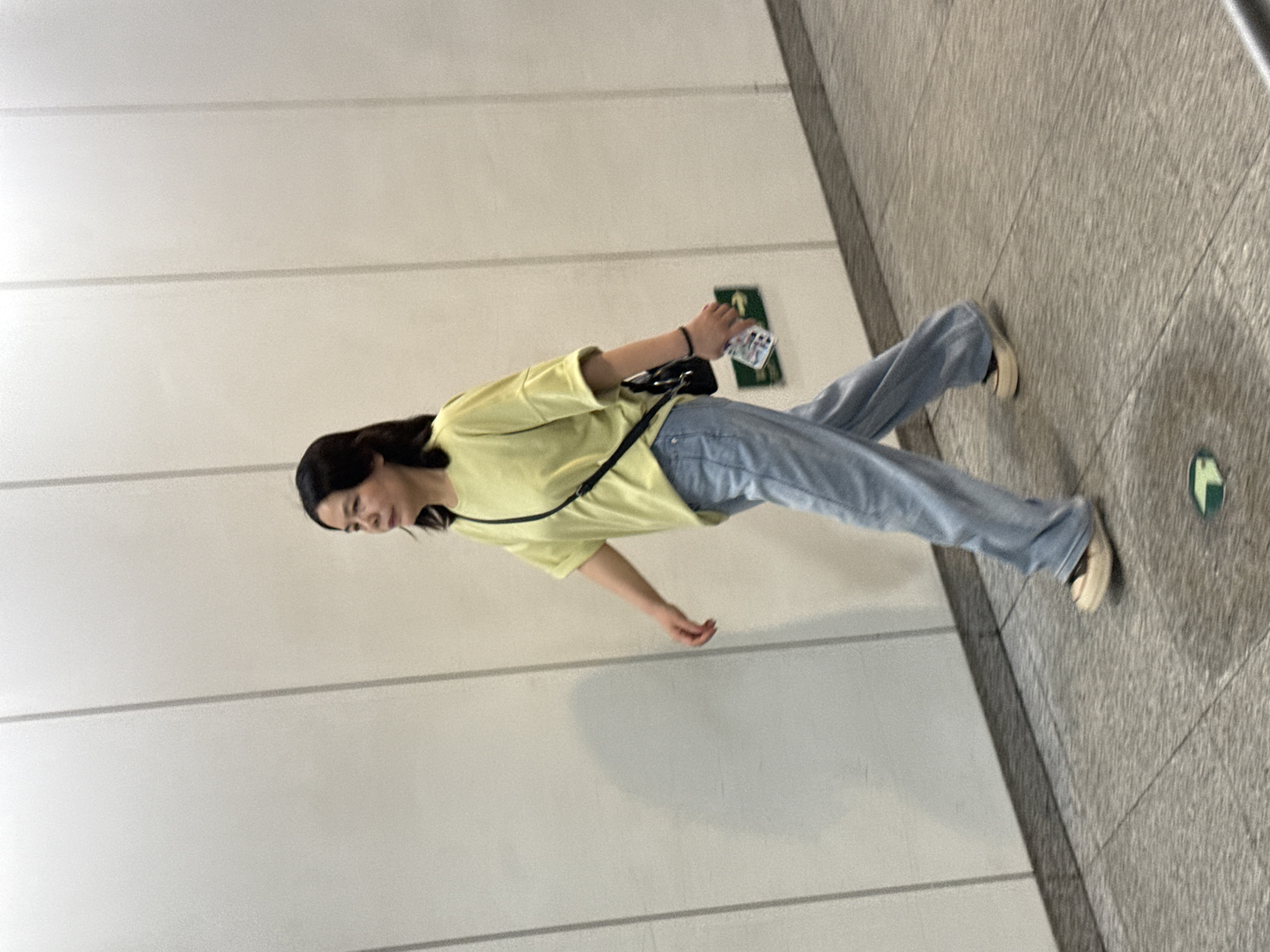 The image, taken in what appears to be an indoor public space, captures a young woman in motion. She has long, dark hair and is wearing a light yellow short-sleeved t-shirt, light blue loose-fitting jeans, and light-colored slip-on shoes. A black cross-body bag with a thin strap is visible across her chest. She holds a smartphone with a colorful, possibly cartoon-themed, white case in her right hand.
The woman is depicted mid-stride, walking across a grey, textured tiled floor. To her left is a plain light grey or white wall with subtle horizontal seams or panels. Behind her, attached to this wall, is a green rectangular sign with a white upward-pointing arrow, likely indicating direction or an exit. A small, green, possibly circular or square symbol is also visible on the floor near her right foot.
The setting suggests a modern public interior like a subway station corridor or a shopping mall in Nan'an District, China. The lighting is even and artificial, typical of such indoor environments. Her expression is neutral, and she appears to be walking purposefully. There is no decipherable text on the phone case or the wall sign beyond the white arrow. No specific time of day or weather conditions can be determined from the indoor scene.