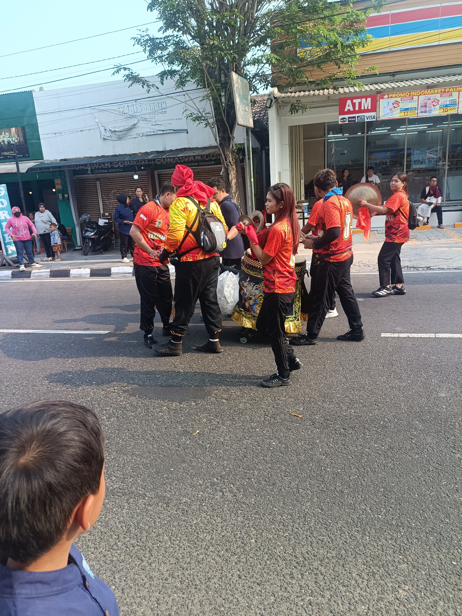 In the city of Sleman, Indonesia, a street procession is taking place along a paved road. Several individuals are dressed in vibrant orange and yellow outfits, some adorned with red headscarves, suggesting a cultural performance or parade. One person is playing a large drum, while another holds cymbals. A trolley-like structure, decorated with intricate patterns and gold trim, is being pushed by participants. On the sidewalk, several onlookers, including children, are observing the event. Behind them, buildings line the street, including a minimarket with an ATM visible. The environment is characterized by a clear sky and trees casting shadows on the road, indicating it is likely daytime. The scene captures a moment of community engagement and cultural expression in a bustling urban setting.
