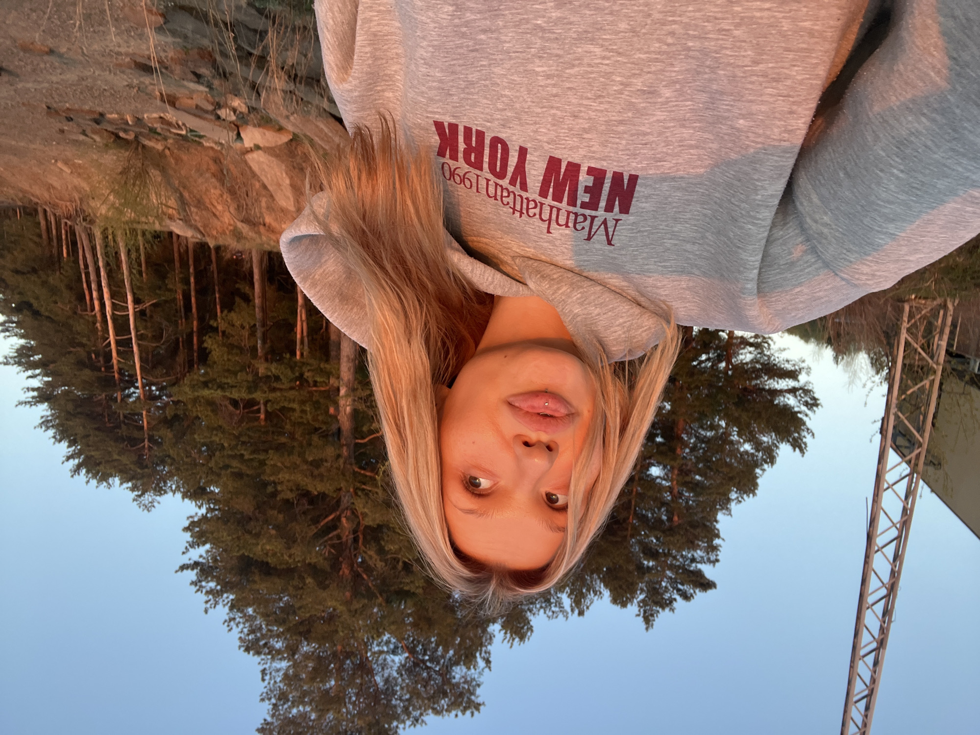 A selfie of a young woman with blonde hair, wearing a grey hoodie with "Manhattan 1990 NEW YORK" printed on it, is taken outdoors. The camera angle is from below, looking up at the woman's face. She has a pierced lip. Behind her, there are evergreen trees and what appears to be a rocky outcrop. A metal tower is also visible in the background. Based on the context of the city of Chaikovsky, Russia, the location is likely a wooded area or park within or near the city, although specific landmark identification is not possible from the image alone.