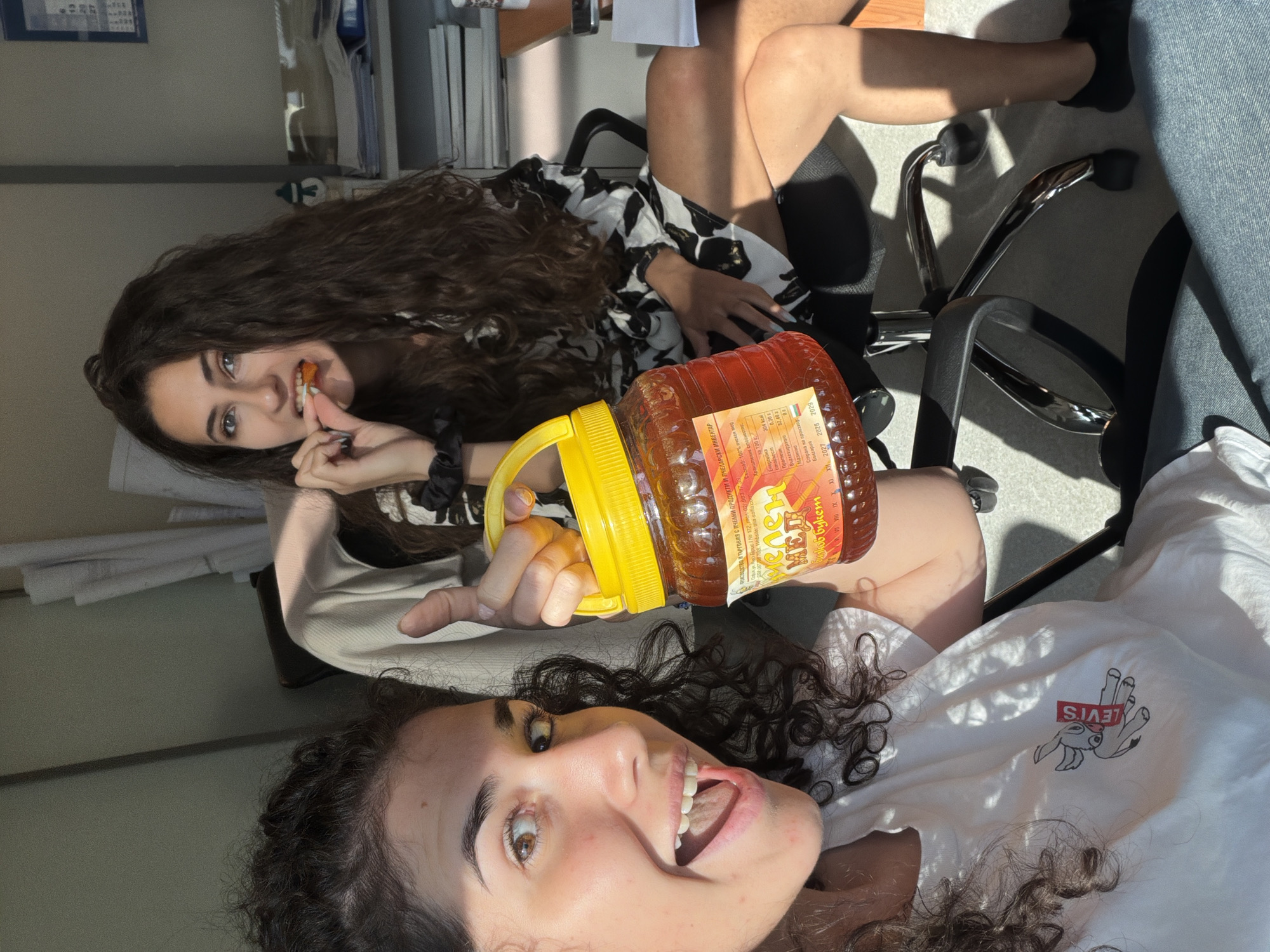 Two young women are in an office environment. The woman in the foreground is on the left, looking at the camera with an open mouth and a wide smile. She is holding a large, clear plastic jar of honey with a yellow lid and handle. The label on the jar is partially visible and appears to be in Cyrillic script. She is wearing a white t-shirt with a graphic of a donkey. The woman in the background is on the right, seated, and is eating something orange from her fingers. She has long, dark, curly hair and is wearing a black and white patterned dress. The office appears to be somewhat cluttered with papers and office supplies visible in the background. Sunlight is streaming in from the left, casting strong shadows. The scene is likely a candid moment captured in an office in Sofia, Bulgaria.
