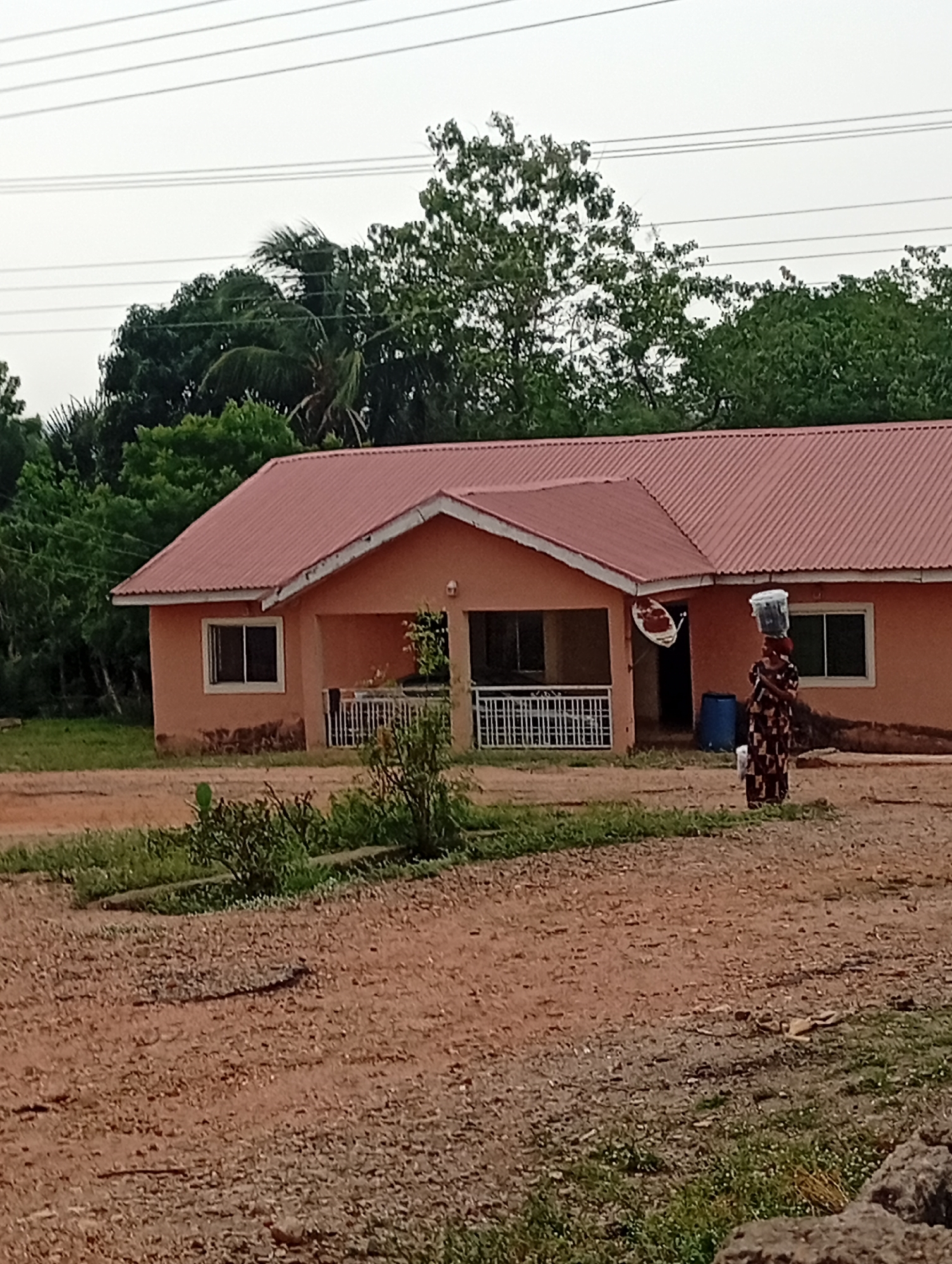 A woman wearing a patterned brown dress is walking toward the camera, balancing a white container on her head. The woman is on a dirt path in front of a single-story peach-colored building with a rust-colored corrugated metal roof. The building has two windows and a porch with a white railing. Green trees are in the background and power lines stretch across the sky. The location appears to be in Adankolo, Nigeria, based on the context provided.