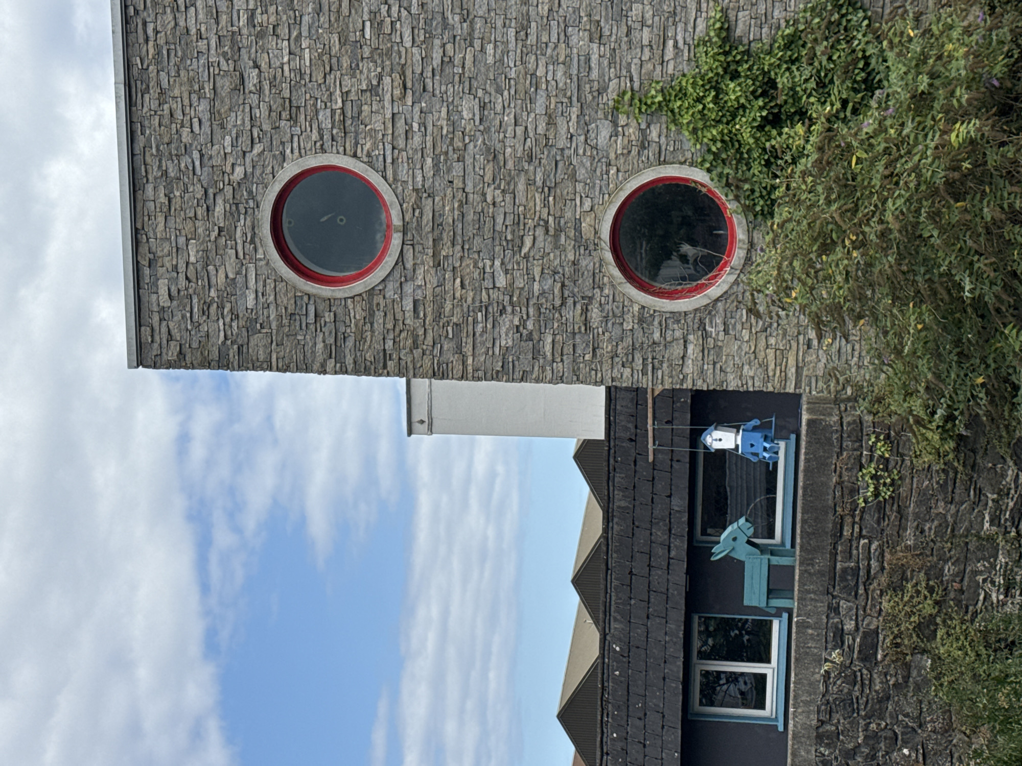 The image shows a modern building with a stone facade and two circular windows with red frames. The building is located in Waterford, Ireland. In the foreground, there is a wall made of rough stones, and behind it, a lower building with a dark roof and two white windows. To the left of the windows, there is a blue wooden donkey figure, and to the right, a blue figure of a person on a swing. Lush green bushes and ivy grow around the base and side of the stone building. The sky is blue with scattered white clouds.