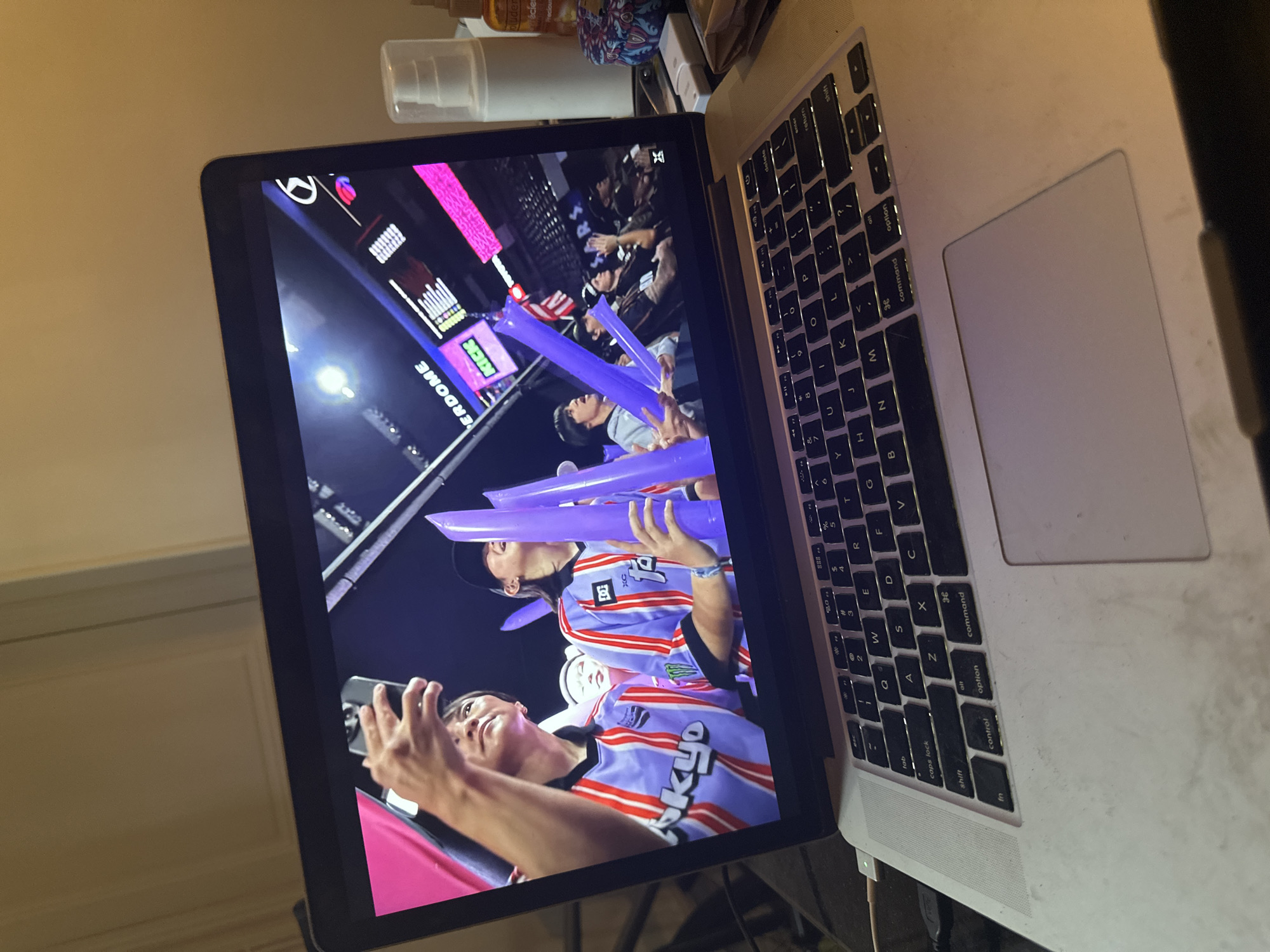 This is a close-up, eye-level, medium shot of a laptop displaying a video of a sporting event, likely basketball, shown on its screen. The laptop is open and positioned on a desk or table. The screen shows a crowd of people watching the game, some holding purple inflatable noisemakers. Two individuals in the foreground on the screen are wearing purple and white striped jerseys. The background on the screen shows a stadium with bright lights and advertisements, including one that reads "KICK" and another partially visible "ERDOME." The laptop itself is silver, with a black keyboard and a silver trackpad. The overall scene is dimly lit, with the primary light source coming from the laptop screen. The right side of the laptop shows a white spray bottle and a small bottle of hair oil. The laptop is not in Paris, France.