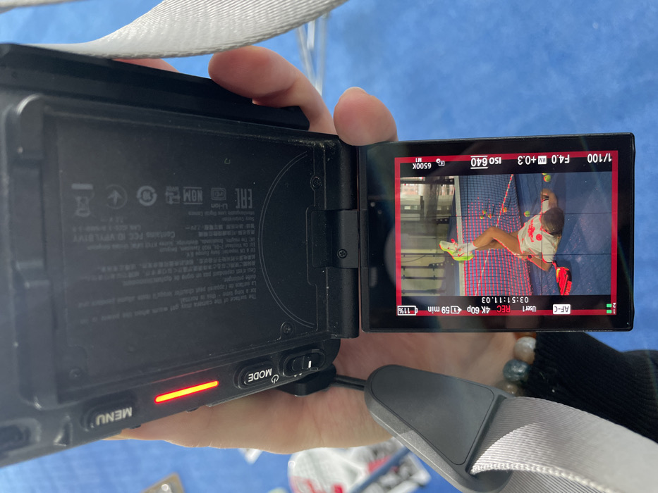 The image displays a close-up, top-down view of a black digital camera, identified as a Sony ZV-E10 by its FCC ID VP7LB1Y visible on the open back panel. A right hand, adorned with a multi-colored beaded bracelet on the wrist, is gripping the camera. The camera's articulated LCD screen is flipped out and angled towards the viewer. A red horizontal indicator light is illuminated to the right of the "MENU" button on the camera's left side. The background behind the camera is a blurred blue surface with indistinct white objects.
The camera's screen displays a high-definition video feed or recorded footage in portrait orientation. Technical overlays indicate "4K 60p" resolution, "AF-C" focus mode, a battery charge of "11%" with "1h 59 min" recording time remaining, and a timecode "03:51:11.03". Exposure settings show "ISO 640", "F4.0", "+0.3" exposure compensation, and "1/100" shutter speed. The white balance is set to "6500K". The footage depicts a person, appearing female, lying on their back on a blue padel court. The person wears a white athletic shirt with red patterns, dark shorts, and bright yellow-green athletic shoes. One leg is bent at the knee and raised. A red padel racket rests on the court beside the person. Several yellow-green padel balls are scattered around the individual. The court features red boundary lines and a white net. The background of the court shows an indoor sports facility with grey structural elements.
This scene is located at the Padel Club Ekaterinburg, situated at Ulitsa Malysheva, 74, Yekaterinburg, Russia.
