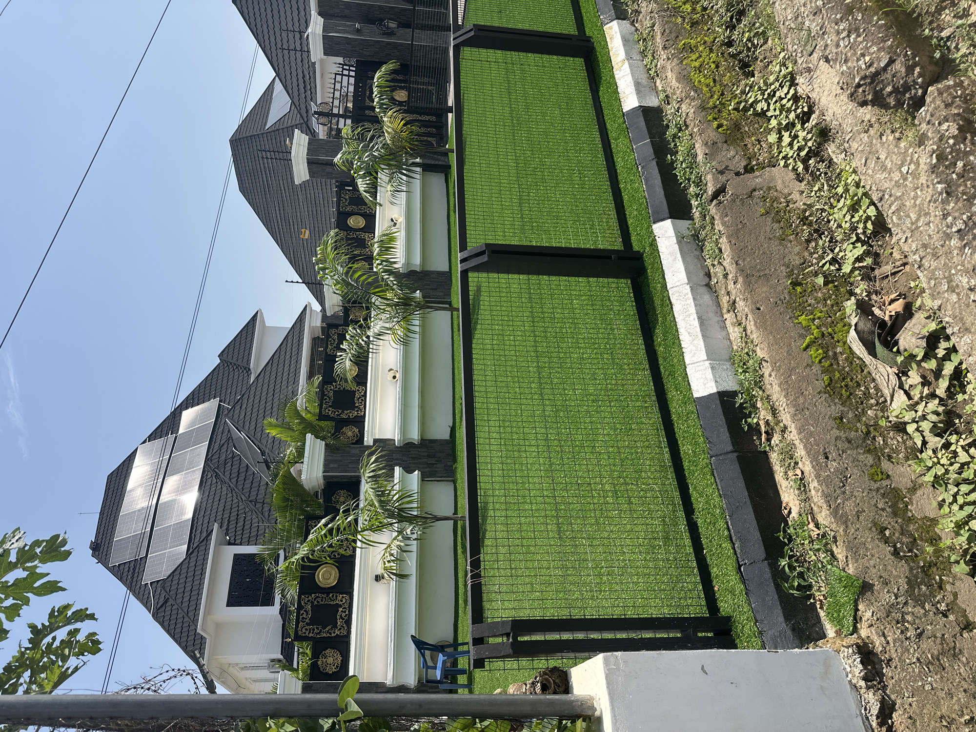 The image depicts a modern residential property in Abuja, Nigeria, featuring a multi-gabled roof adorned with solar panels. The house has a white facade with decorative black and gold embellishments along the top of a low wall. A black metal fence encloses a neatly manicured lawn made of artificial turf. Small palm trees are planted within the fenced area. A blue plastic chair is visible on the artificial lawn. The property is bordered by a concrete curb painted with alternating black and white stripes, and beyond that, a concrete drainage ditch with overgrown vegetation and some moss. The sky is clear blue, and power lines are visible crossing the upper portion of the image. The overall impression is of a well-maintained, contemporary home in an urban setting.