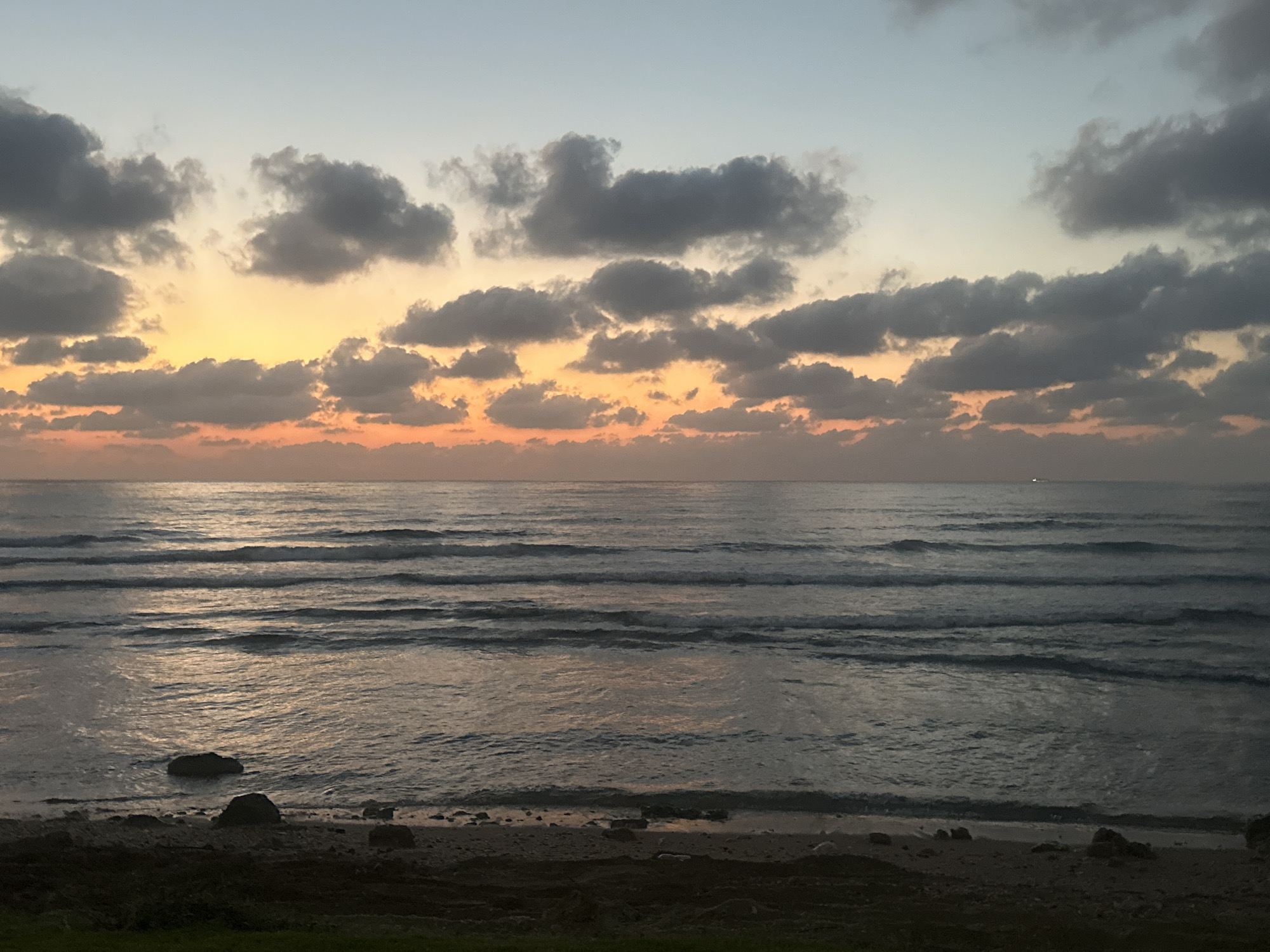 A serene beach scene at dusk in Haifa, Israel, showcases the calming effect of nature. The sky is a canvas of soft hues, transitioning from a pale blue at the top to warm oranges and pinks closer to the horizon, painted with streaks of scattered gray clouds. The ocean, a vast expanse of deep blue-gray water, mirrors the sky's colors with gentle waves rolling towards the shore. The beach itself is a mixture of sand and rocks, with larger stones scattered along the waterline. A few dark rocks are visible in the shallow water, adding texture to the scene. In the distance, a faint light suggests the presence of a boat on the water. The foreground is a dark, uncultivated strip of land, likely the edge of a park or vegetation, with a hint of green grass visible at the very bottom. The overall impression is one of tranquility and natural beauty, capturing a peaceful moment as day turns into night.