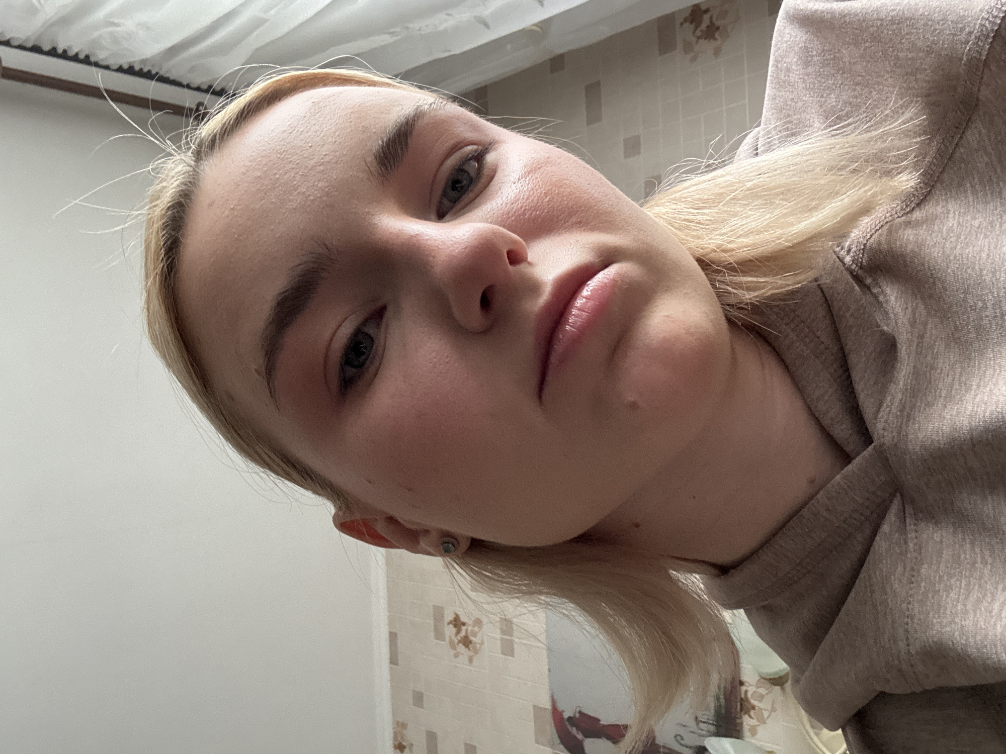 A close-up, low-angle, indoor shot shows a young Caucasian woman with blonde hair and fair skin. She is looking directly at the camera with a neutral expression. She is wearing a beige hooded sweatshirt. The background consists of a wall with light-colored wallpaper featuring a subtle floral pattern, and a white curtain on the right side. The lighting appears to be natural, coming from an overhead source. The image appears to have been taken in Sinyavino, Russia, based on the provided context.