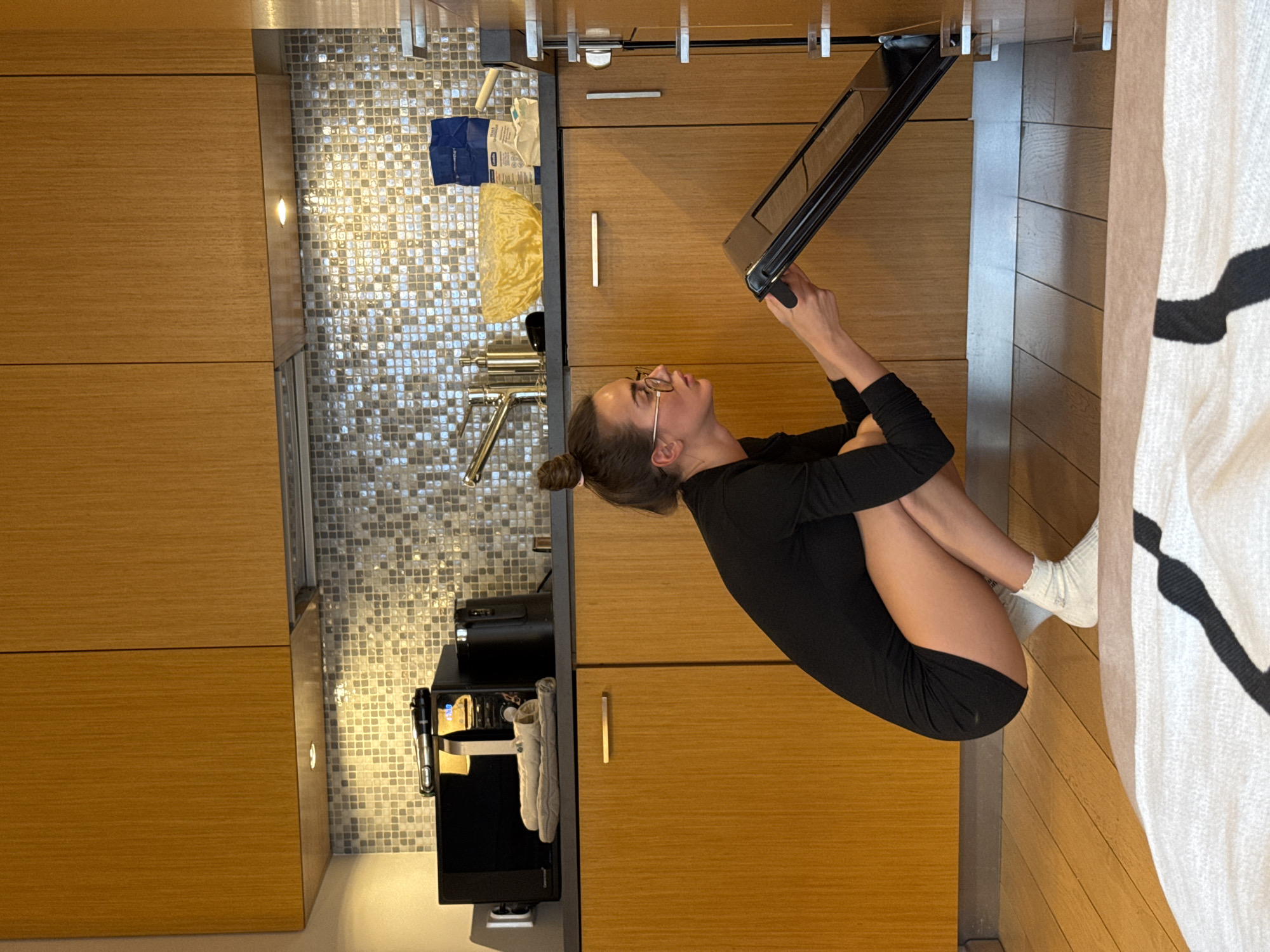 A woman in a black dress and white socks is crouched on a wooden floor in a modern kitchen, possibly in Warsaw, Poland. She is looking up and holding a black frame. The kitchen features wooden cabinets, a mosaic backsplash, a coffee machine, and a sink. A patterned rug lies on the floor to the right of the woman.